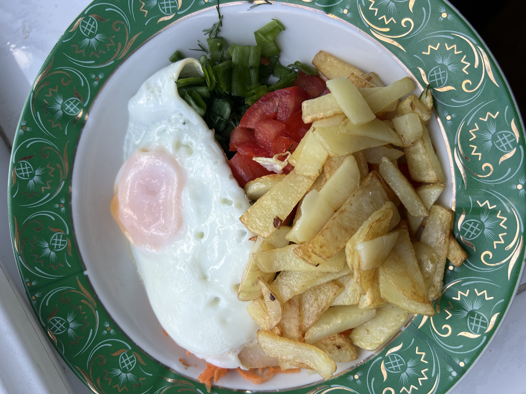 A close-up, top-down photograph captures a freshly prepared meal served in a decorative bowl. The main components of the dish are a sunny-side-up fried egg with a firm white and a soft, translucent pinkish-orange yolk, placed on the left side of the bowl. Adjacent to the egg is a generous portion of golden-brown, thick-cut fried potatoes or fries, some exhibiting slight crisping and browning.
Between the egg and the fries, a cluster of diced red tomatoes is visible. Above the egg and tomatoes, a heap of freshly chopped green onions or chives, along with finer strands of dill, adds a vibrant green. Beneath the egg and other ingredients, a thin layer of finely grated orange vegetable, likely carrots, is subtly present.
The meal is presented in a white bowl featuring a wide, ornate emerald green rim. This rim is intricately decorated with golden scrollwork patterns, intertwined with white and gold stylized motifs that resemble pineapples or thistles, each with a distinctive crosshatch design. Zigzag lines in white and gold further adorn the rim.
The lighting is bright and natural, suggesting a daytime setting. The background is softly blurred, showing indistinct lighter and darker tones, possibly indicating a table or surface near a window. There are no people visible, and no text is present in the image. The meal appears fresh and ready for consumption.