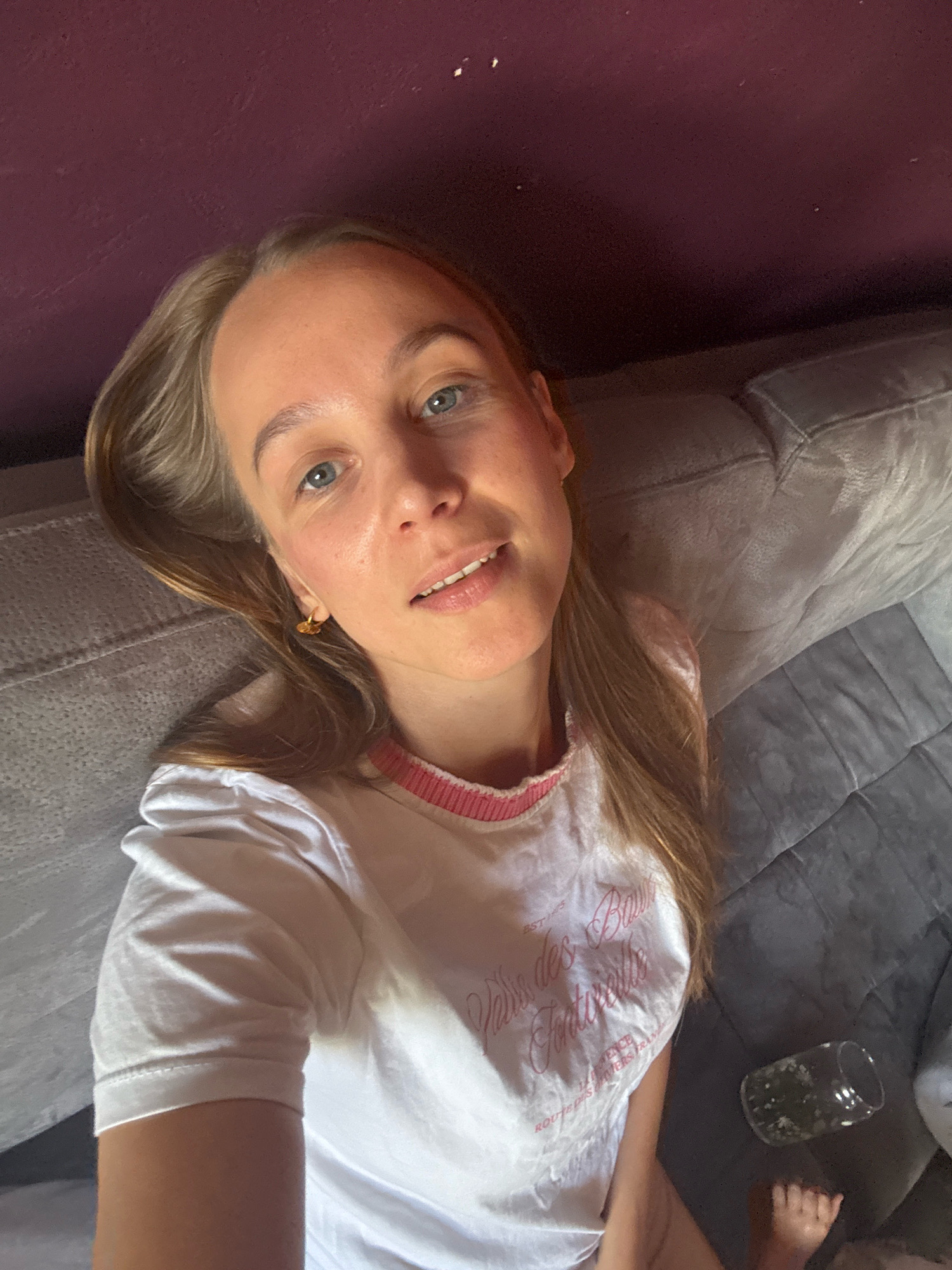 A young woman is lying down on a grey couch, taking a selfie. She has long, blonde hair and blue eyes, and is wearing a white t-shirt with pink trim. A small gold earring is visible in her left ear. The text on her shirt reads "EST 1785 Vains des Fontenelles" and "12 AVENCE ROUTE DES RIVIERS TRAM". A glass containing a green liquid and ice is on the couch to her right, near her feet. The background includes a dark purple wall and the textured surface of the couch. The lighting suggests it is daytime.