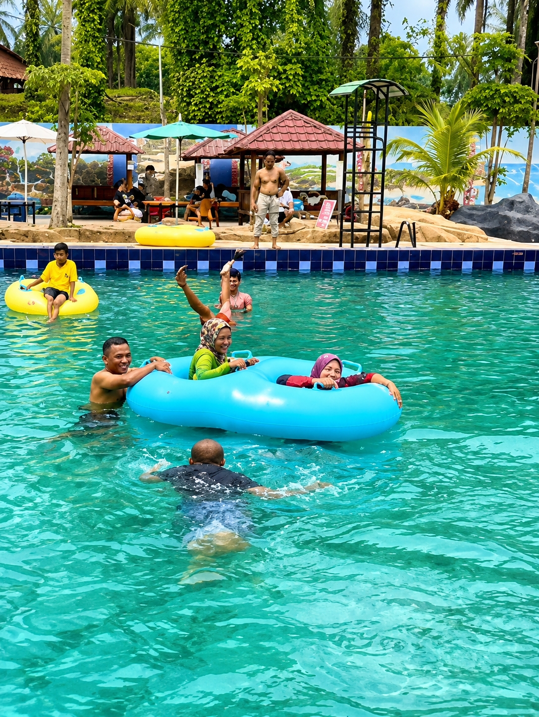 In the city of Eampat Kwalamencirim, Indonesia, this image captures a vibrant scene at an outdoor water park. The turquoise water of the swimming pool is filled with people enjoying various water activities. In the foreground, a group of individuals are interacting within a large, blue inflatable tube. One person is paddling in the water, facing away from the camera. Further back, a young boy in a yellow shirt sits on a yellow inflatable ring. The background reveals the park's amenities, including several small, thatched-roof huts, lounge chairs, and umbrellas. Lush green trees and palm fronds surround the area, suggesting a tropical climate. The overall atmosphere is one of recreation and leisure, with people of different ages engaging in water-based fun.