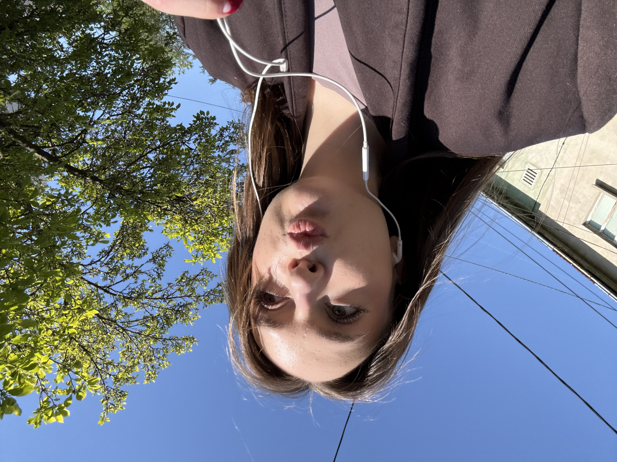 The image is an inverted, low-angle self-portrait taken outdoors on a clear, sunny day in Kragujevac, Serbia. The primary subject is a person, appearing female, with long brown hair, occupying the central and lower portions of the frame, presented upside down. She is wearing a dark brown jacket or coat and white wired earphones, which are visible around her neck and shoulders. Her face, seen from below and inverted, features a slight pout and her eyes are directed upwards. The strong sunlight illuminates her face, causing some shine on her forehead, and creates defined shadows on her clothing and neck.
The background consists of a vibrant blue sky, suggesting bright daylight. To the left and top (when viewed upright), lush green foliage from a tree is visible, brightly lit by the sun. In the upper right section of the image (when viewed upright), parts of a building structure with a vent and a window are discernible against the sky. Black power lines crisscross the blue sky in the right portion of the frame, indicating an urban or suburban setting.
The activity captured is a self-portrait, likely a selfie, taken from an unusual perspective, possibly with the camera held below the subject and then inverted. The person appears to be casually posing. There is no visible text in the image. The bright sun and clear sky indicate pleasant weather conditions.