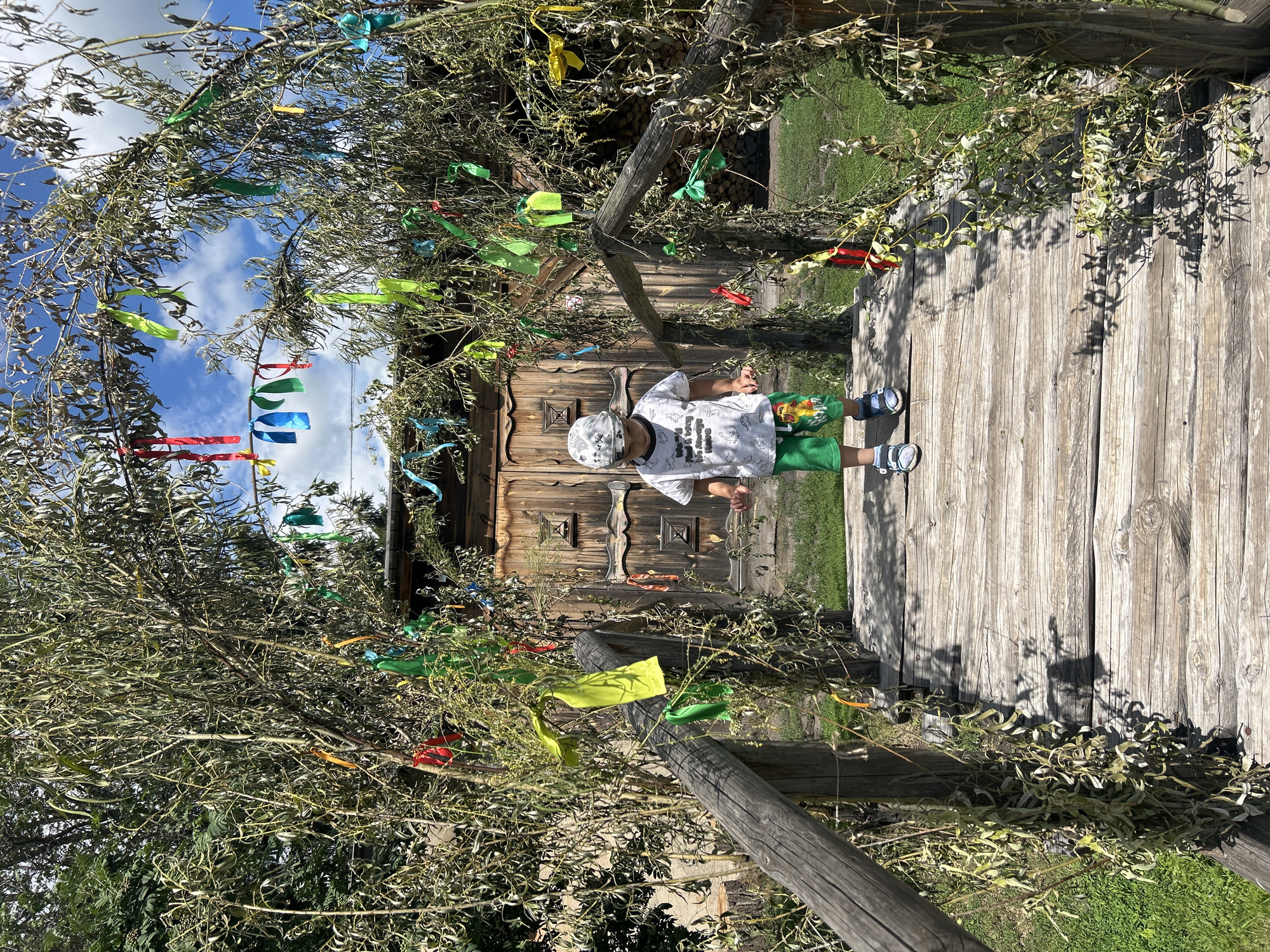 A young boy stands on a wooden bridge adorned with colorful ribbons and dried branches. He is wearing a light-colored t-shirt with a graphic print, green shorts, a patterned cap, and sandals. The bridge leads towards a rustic wooden building with a double door. The scene is set outdoors, likely in a village or rural area, given the natural foliage and the building's construction. The background shows a grassy area and a blue sky with scattered clouds. The overall impression is one of a festive or decorative outdoor setting, possibly a village festival or a specially decorated area in Bolsherechye, Russia.