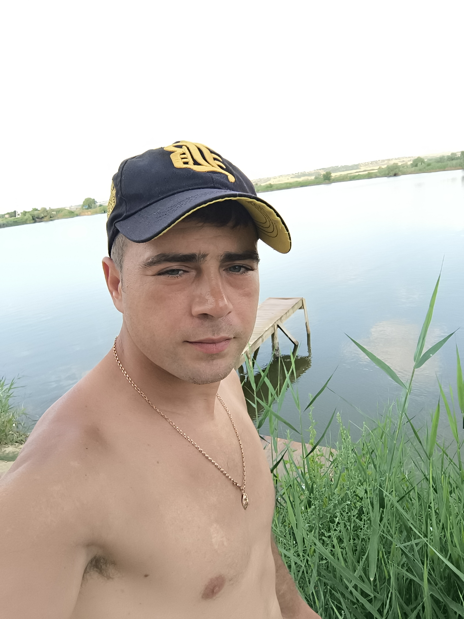 A man wearing a dark blue baseball cap with a yellow emblem and a gold chain necklace stands shirtless in front of a body of water. The man is looking directly at the camera. To his right, there is tall green grass and a wooden dock extending into the water. The background shows a distant shoreline with sparse vegetation and a pale sky. The setting appears to be a natural outdoor environment. There is no indication that the image was taken in Ciumai, Moldova.