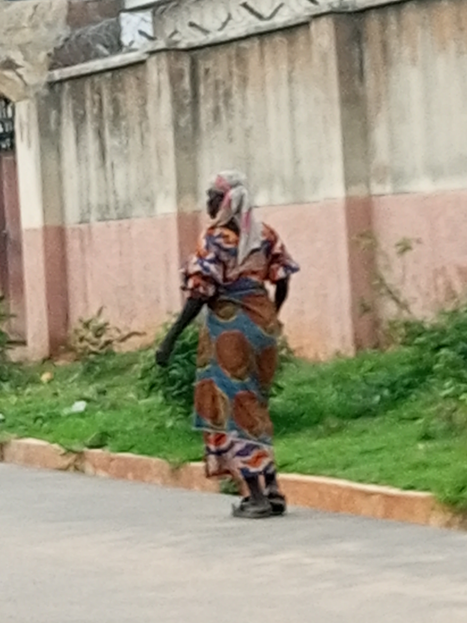 A person is walking away from the camera on a paved surface next to a grassy area. The person is wearing a colorful patterned dress and a headscarf. Behind the person is a weathered wall with some decorative elements at the top, painted in shades of beige and pink. Green vegetation grows along the base of the wall. The image is taken outdoors in what appears to be a city setting.