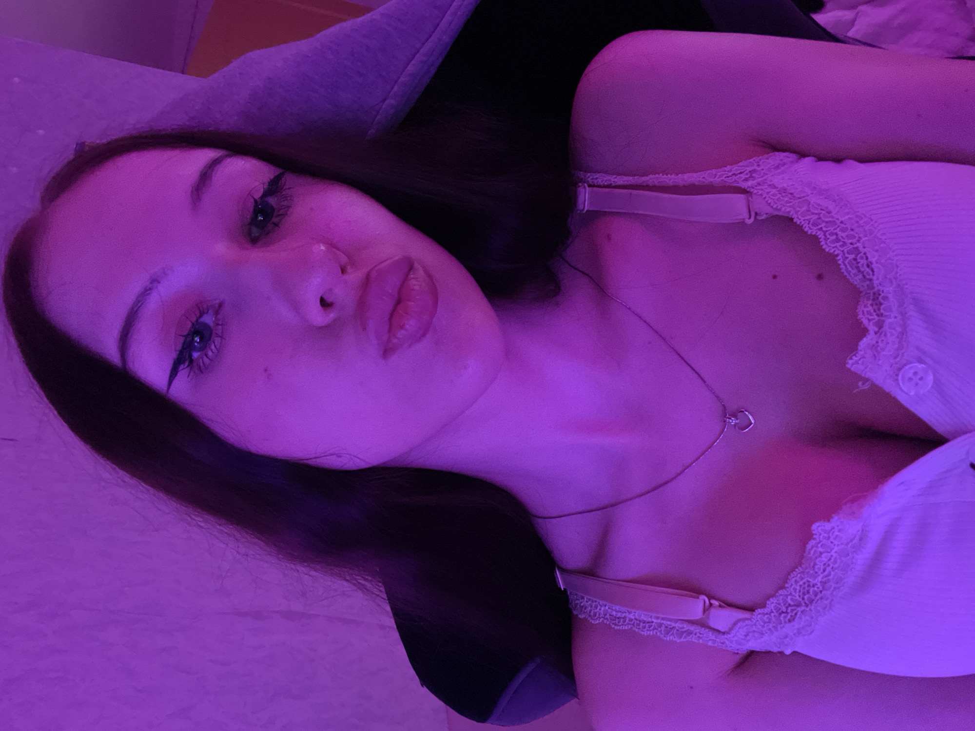 The image shows a close-up of a young woman from the chest up, posed in front of a textured, purple-lit background. She has long, dark hair, and her face is turned towards the camera. She is wearing a light-colored camisole with lace trim and a delicate necklace with a heart-shaped pendant. The lighting is atmospheric and predominantly purple, casting shadows and highlighting her features. The background appears to be a wall or fabric surface. There is no discernible location in the city of Votkinsk, Russia, depicted in this image.