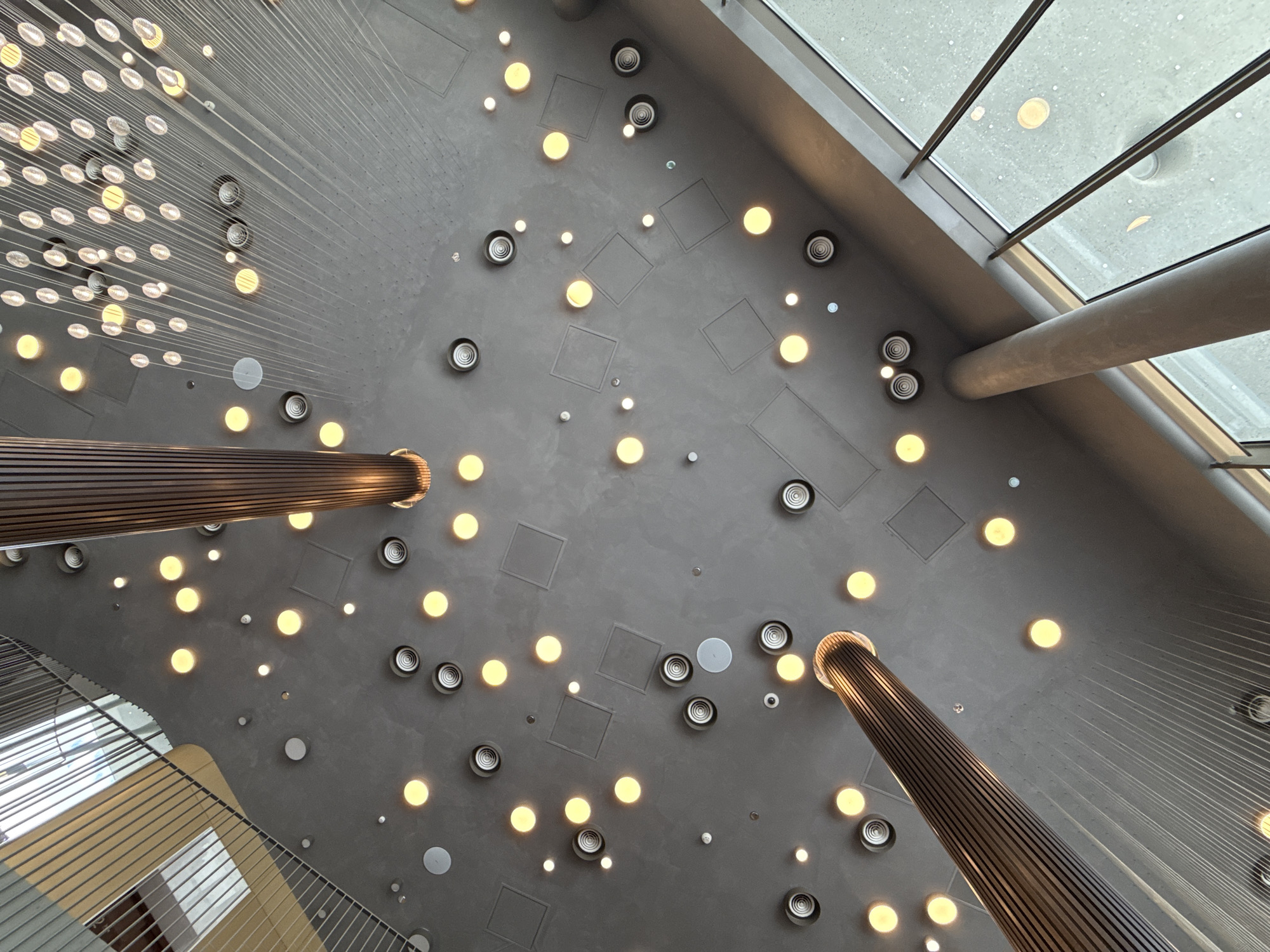 The image shows a low-angle view of a modern interior space, likely a public building or atrium. The dominant feature is the ceiling, which is a dark, matte gray material. It is sparsely populated with various circular elements. Several large, warm yellow circular lights are evenly distributed across the ceiling, some appearing to be recessed spotlights. Interspersed between these are smaller, white circular lights. Also visible are circular ventilation grates with a spiral pattern and some square ceiling panels.
Two large, cylindrical pillars, clad in dark wood with vertical slats, rise from the floor and extend upwards, disappearing into the ceiling. These pillars are a significant architectural element, adding a sense of scale and verticality to the space.
Thin, white wires or cables are suspended from the ceiling in a dense, arcing pattern, creating a visually complex, almost net-like effect over a large portion of the upper right area of the image. These cables are illuminated by numerous small, circular lights, possibly part of a decorative installation or a specialized lighting system, giving the impression of a starry sky or a network of fine threads.
On the left side of the image, a section of a balcony or walkway with a metal railing and yellow-toned panels is visible, suggesting multiple levels within the building.
On the right side, large windows are present, revealing an overcast or rainy outdoor environment, with blurred circular reflections of lights on the glass.
The scene is devoid of any people or direct human interaction. The overall impression is one of contemporary design with a focus on lighting and architectural form, creating a sophisticated and possibly awe-inspiring atmosphere.
The request for information regarding the location in "Голубинское сельское поселение, Ukraine" cannot be fulfilled as there is no visual information in the image to support or refute this specific location.