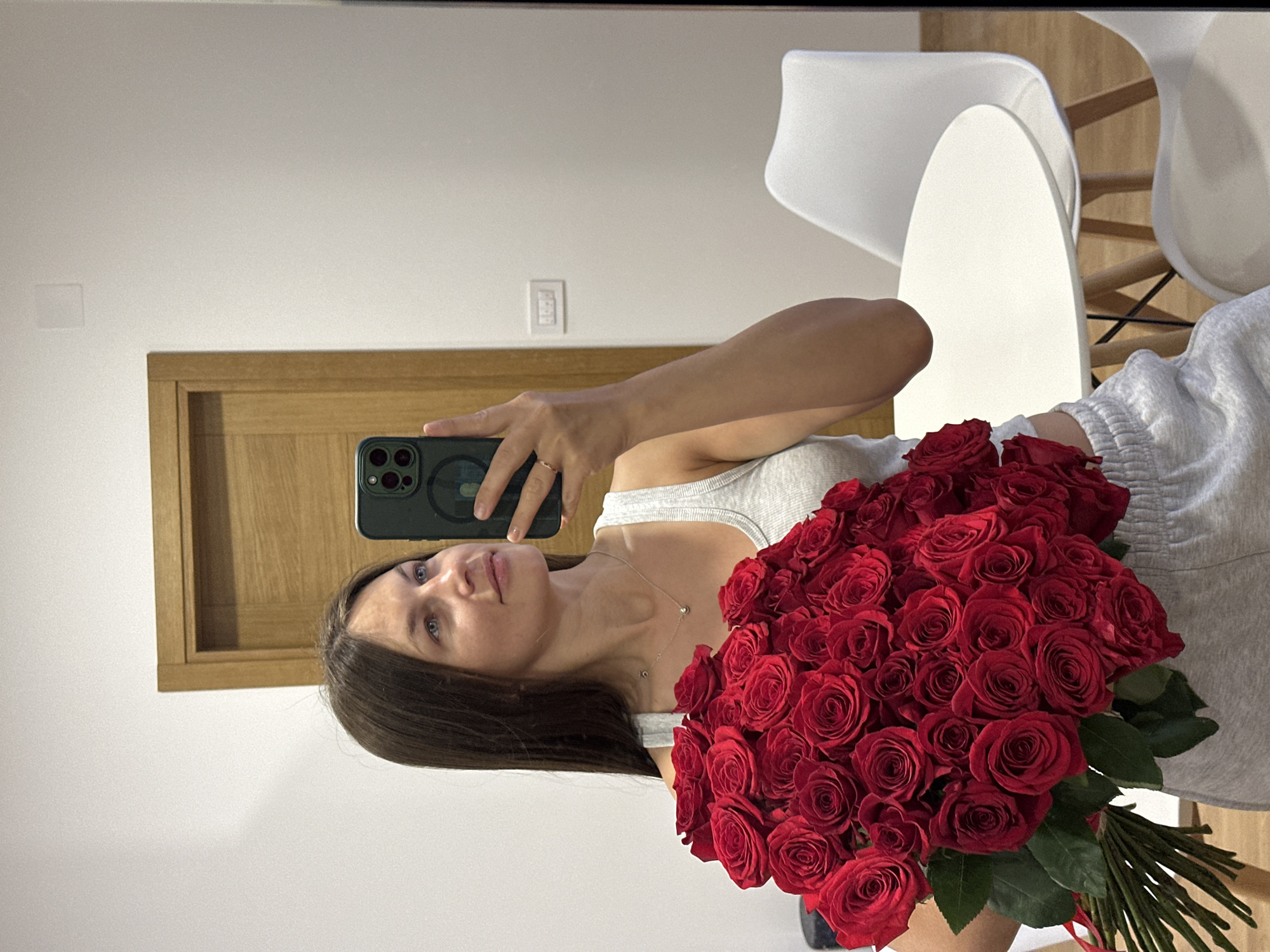 A woman with long brown hair holds a large bouquet of red roses in front of her while taking a selfie with a phone. She wears a light grey tank top and light grey sweatpants. She is wearing a ring on her ring finger and a necklace. The background includes a white wall, a light brown door frame, a light switch, and two white chairs next to a white table. The picture is taken in a room indoors. The location is Donji Pobori, Montenegro.