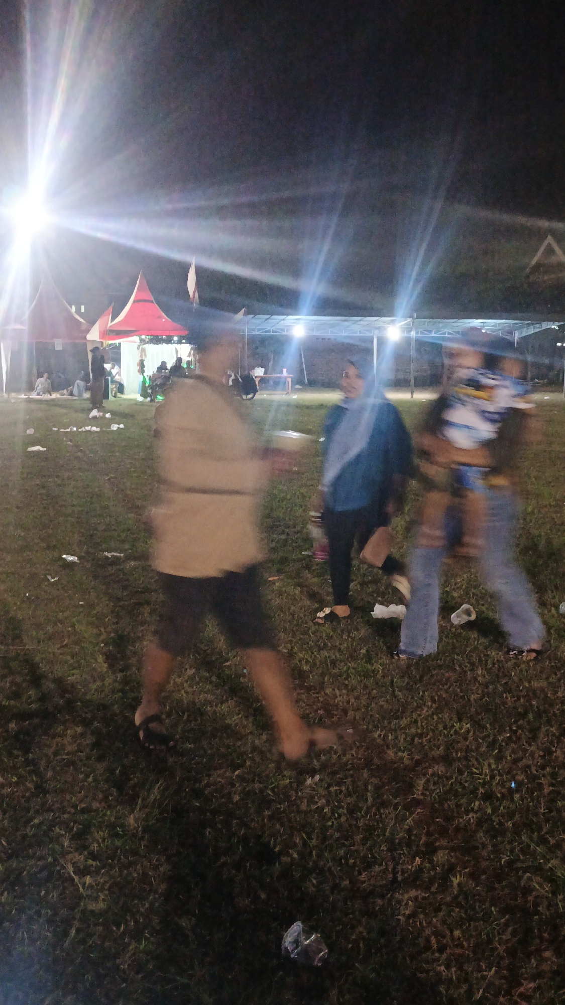 A nighttime outdoor scene depicts an event with people and tents. In the foreground, a person in a light-colored long-sleeved shirt and dark shorts is blurred, appearing to walk to the left. To the right of this individual, a person wearing a blue hijab and a dark jacket walks toward the foreground. Further to the right, another person, mostly blurred, is walking away, wearing a white and blue patterned top and blue jeans. Several tents are visible in the background, some red and white, and a stage-like structure with lights is set up. The ground is covered in grass and scattered litter, including plastic cups. The overall lighting suggests the event is illuminated by artificial lights and possibly flash photography, causing some motion blur in the figures. The location is identified as Ketemasdungus, Indonesia.