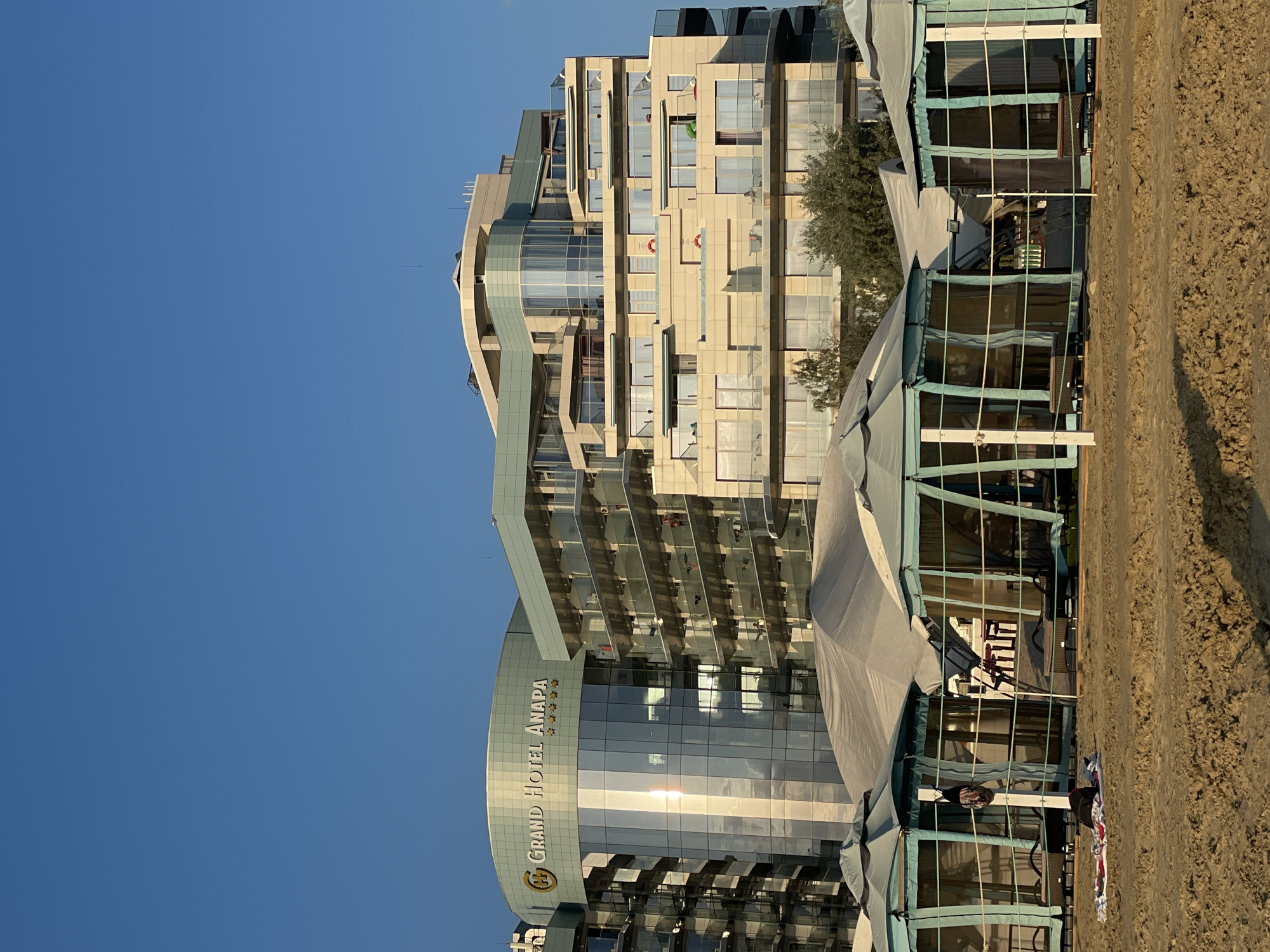 The image shows a modern, multi-story hotel with a curved glass facade and a sandy beach in the foreground. The hotel is branded "Grand Hotel Anapa" with a logo and several stars visible on the glass. A large, light-colored awning structure extends from the hotel towards the beach, with tables and chairs set up underneath, suggesting an outdoor dining or lounge area. Some parts of the awning are opaque, while others have transparent panels, allowing views into the space. The beach is sandy and appears to be relatively empty. The sky is clear and blue, indicating good weather, and the lighting suggests it is late afternoon or early evening, with the sun casting long shadows and reflecting off the hotel's windows. A lone tree is visible near the hotel. The overall scene depicts a resort setting on a sunny day.