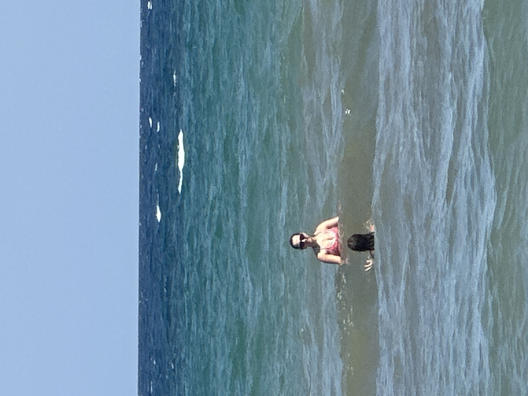 Two individuals are in the ocean. A woman wearing a pink bikini and sunglasses is standing in the water. In front of her, a person with dark hair is partially submerged in the water. The ocean is choppy with visible waves. The sky is clear and blue.