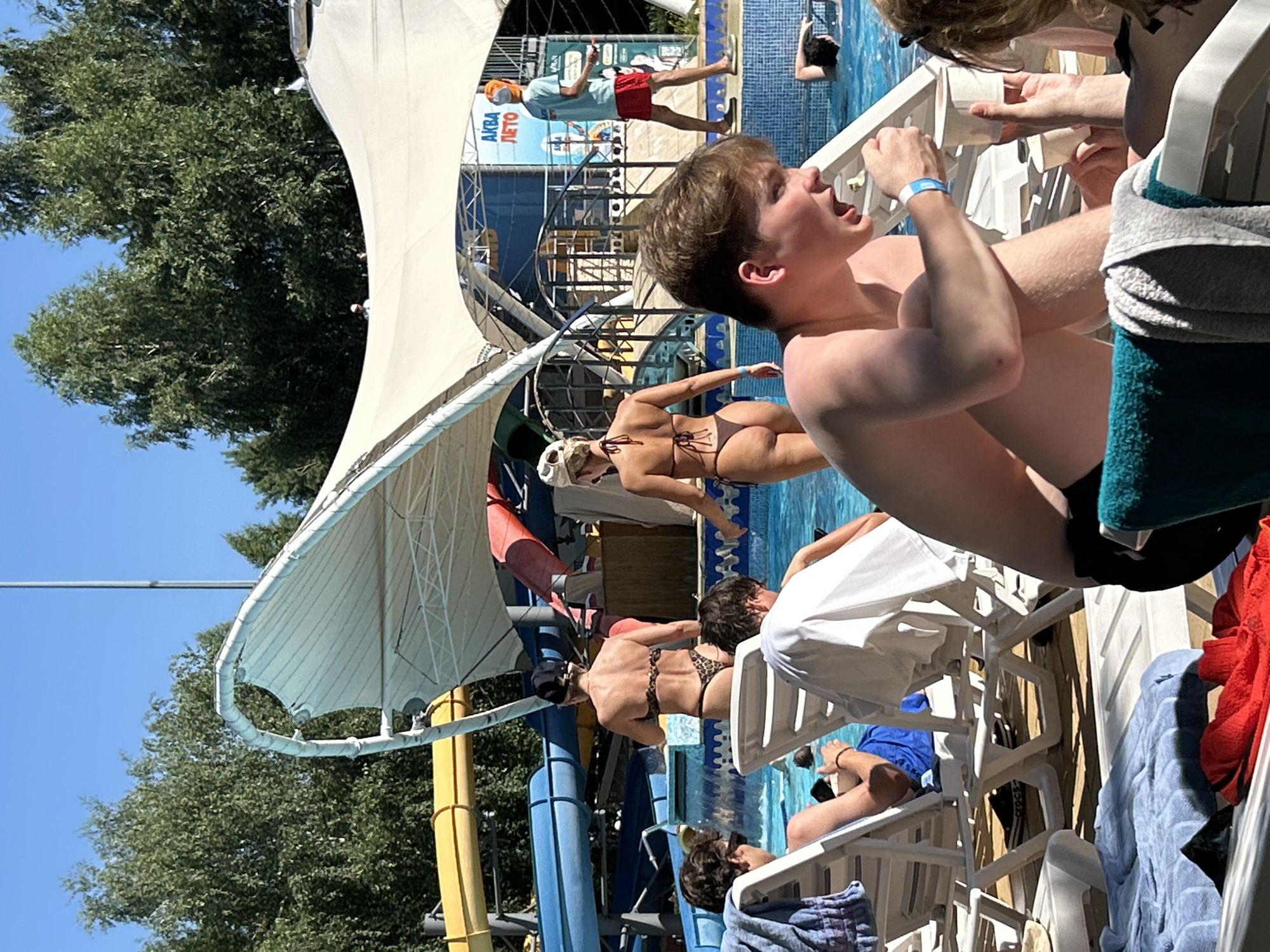 The image depicts a sunny outdoor scene at the Aqua-Leto water park in Togliatti, Russia. In the foreground, a young man wearing black swim trunks is sitting in a lounge chair, holding a white cup and appearing to be speaking animatedly. To his left, another young man is reclined in a lounge chair, partially obscured, with a blue towel draped over his legs. In the middle ground, several individuals are visible around a swimming pool. A woman in a leopard-print bikini walks away from the camera, while another woman in a similar bikini walks towards the right. Further back, a man in a blue t-shirt and red shorts walks past a sign with Cyrillic lettering. Water slides, one yellow and blue, are visible in the background. Trees and a white canopy structure are also part of the environment. The overall atmosphere suggests a recreational setting on a warm day.