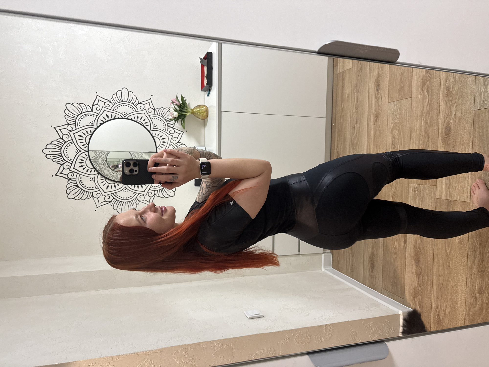 A woman with long red hair is taking a picture of herself in a mirror. She is wearing a black athletic outfit. The mirror reflects a wall with a mandala design, a white cabinet, and a vase of flowers. The floor is wood. The location is likely an interior setting, such as a home or apartment. The image does not contain any information to confirm it is Rostov-on-Don, Russia.