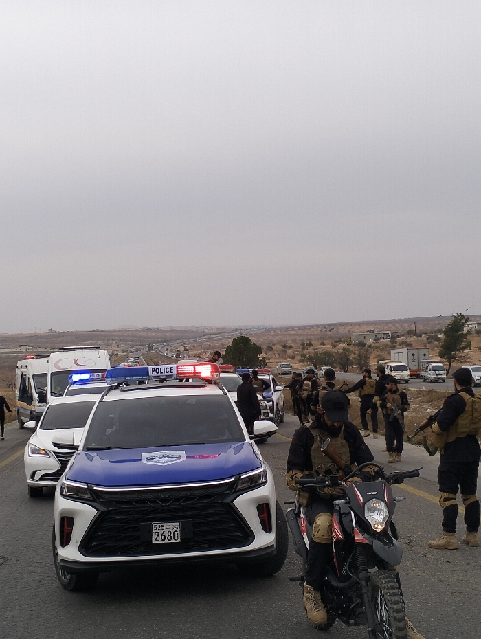 A line of police cars and other vehicles, including an ambulance, is on a road. Several police officers in tactical gear, some holding rifles, are standing beside the vehicles. In the foreground, an officer sits on a motorcycle. The sky is overcast, and the landscape appears arid with distant buildings.