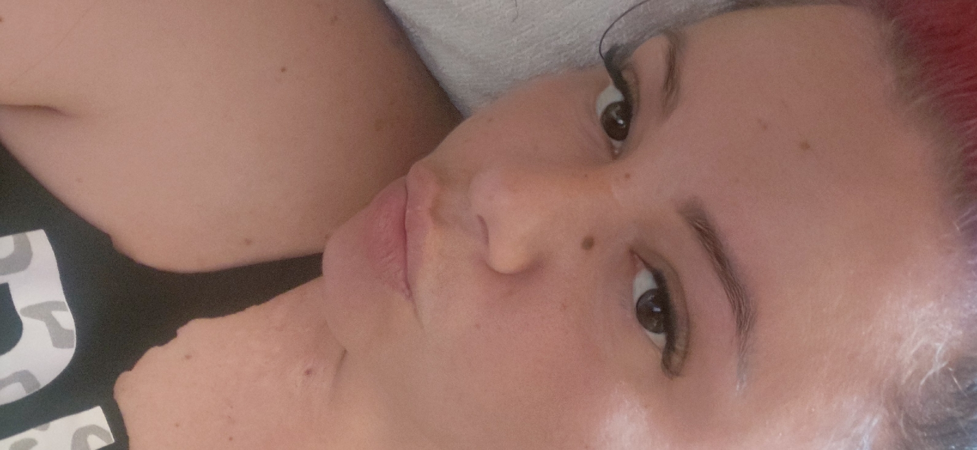 The media file is a close-up, sideways shot of a person's face, specifically focusing on their eyes, nose, mouth, and parts of their neck and shoulder. The person has fair skin with a few small brown moles visible on their face and neck. Their eyebrows are dark and well-defined. The person is wearing makeup, including eyeliner and mascara. Their hair appears to be dark with streaks of bright pink or red at the top.
The scene is lit by artificial light, suggesting an indoor setting. The background is blurry and out of focus, but appears to be a light-colored surface, possibly a bed or sofa. The person is lying down or reclined, looking towards the camera with a neutral expression. There is no discernible activity or event taking place, beyond the person posing for the photo.
Visible text on the person's clothing is partially obscured, but appears to be a large, bold font with a pattern within the letters, possibly a leopard print design. The provided location context is Marynivka, Ukraine, but there are no visual cues in the image to confirm this location. The time of day and weather are not discernible from the image.
