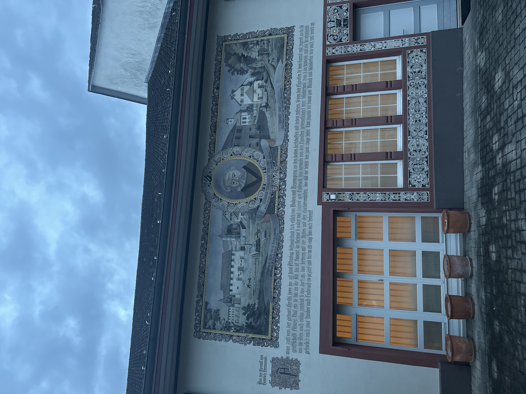 The image displays the side of a building, likely in Embrach, Switzerland, featuring a large historical mural and multiple windows. The scene is set against an overcast, cloudy sky.
The central subject is a detailed mural composed of a portrait and historical landscape scenes, framed by ornate borders. At the bottom left, a circular portrait depicts a man identified as Heinrich Brennwald (1478-1551), described as the "letzter Probst" (last provost) of an "Amtshaus Zehntenscheune" (customs house/tithe barn). Above this portrait, a scene labeled "Embrach Mitte des 15. Jabrhunderts" (Embrach mid-15th century) illustrates a village landscape with buildings. Another scene, "Embrach 1800," shows a similar but possibly evolved view of the locality.
Adjacent to the painted scenes, a lengthy German text from "Aus der Thronif son Heinrich Brennwald" (From the chronicle of Heinrich Brennwald) describes a historical event from 1444. It recounts how forces from Baden and Regensberg, associated with the Lignoffen (Confederates) and the county of Kyburg, advanced towards Embrach. The village was subsequently burned and plundered, leaving only two houses intact, before the forces moved on to plunder Bülach.
The building itself features several windows with white grilles and decorative frames, some of which exhibit a warm, orange glow from internal lighting. Below a large lower window, four terracotta pots are lined up on a ledge. Ornate, dark gray and white decorative patterns adorn the window surrounds and sections of the wall. The time of day appears to be daylight, under a uniformly cloudy sky.