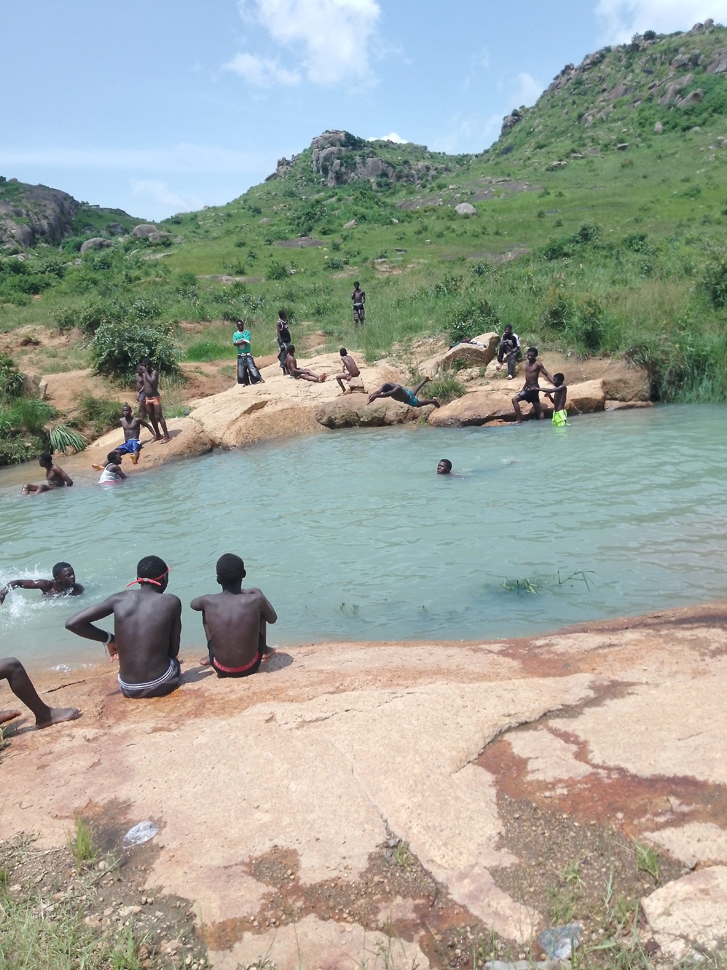 The image captures a group of young men and boys enjoying themselves at a natural swimming spot. The location appears to be rocky with a clear, turquoise body of water. In the foreground, two individuals are seated on a large, tan-colored rock, facing away from the camera, with their backs to the water. One is wearing black shorts with a striped waistband, and the other is wearing black shorts with a red waistband. To their left, a person's legs are visible, suggesting they are either entering or exiting the water.
In the middle ground, several individuals are in the water. One person is in the process of diving into the water from a rock, with their body in mid-air. Another person is swimming, looking towards the shore. Others are wading or sitting on rocks at the water's edge.
The background features a lush, green, hilly landscape with scattered large rocks and vegetation. The sky is bright blue with a few scattered white clouds. The overall impression is one of a recreational outing in a natural, outdoor setting. This scene is consistent with popular informal swimming spots found in and around the city of Jos, Nigeria, known for its hilly terrain and natural water bodies.