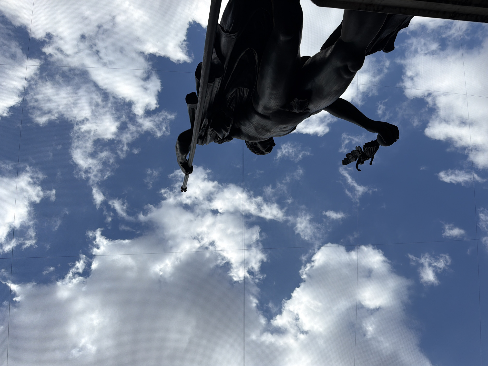 From a low angle, the bronze sculpture of a nude male figure appears against a backdrop of a bright blue sky scattered with white clouds. The figure holds a spear in one hand and what seems to be a winged caduceus in the other. The sculpture is part of the Arco della Pace in Milan, Italy. A faint grid overlay is present, likely from the lens or protective covering used to capture the photograph.