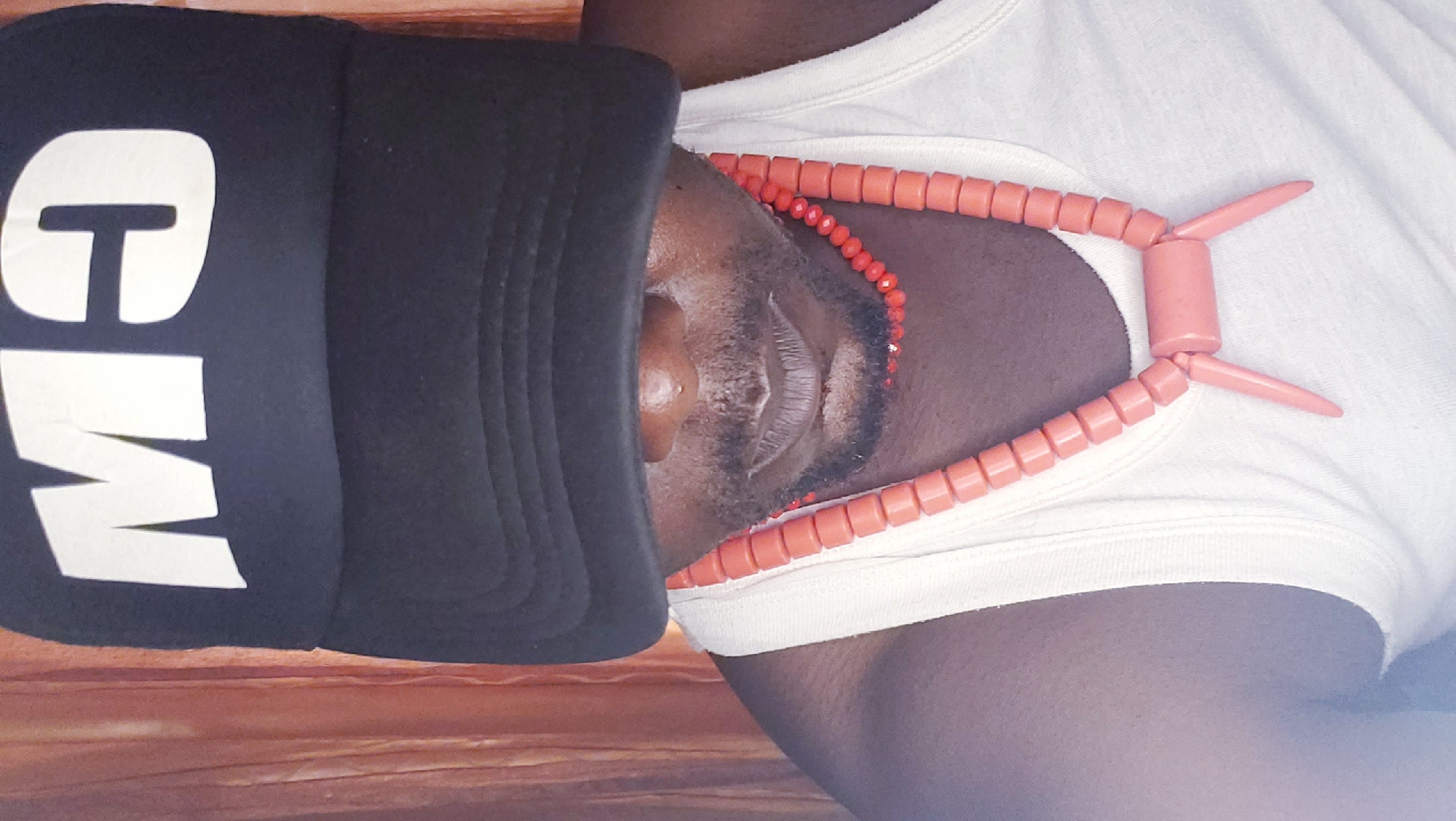 A person wearing a black baseball cap with white lettering that reads "MO" and a white tank top is shown. They are also wearing a coral bead necklace. The person's face is partially obscured by the cap. The background appears to be a light brown curtain. The image is a close-up shot, focusing on the person's upper body. There is no indication of the location being in Narayi, Nigeria.