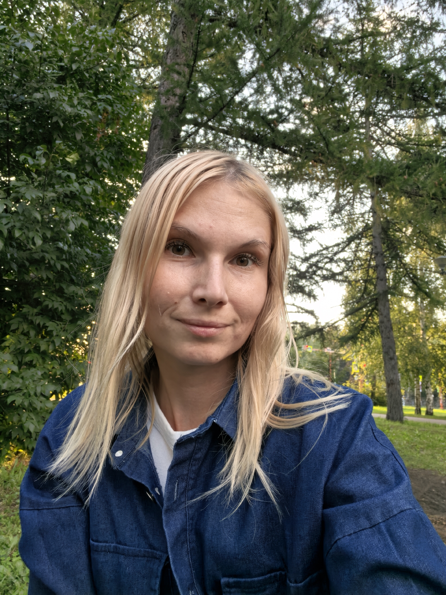 A blonde woman with light brown eyes and fair skin is captured in a selfie outdoors, set against a backdrop of lush green trees and a dappled sky. She is wearing a dark blue denim jacket over a white t-shirt. The woman has shoulder-length blonde hair that is slightly wind-blown. Her expression is neutral with a slight smile. The environment suggests a park or a wooded area within the city of Sterlitamak, Russia. The lighting indicates it is likely late afternoon or early evening, given the warm glow filtering through the trees.