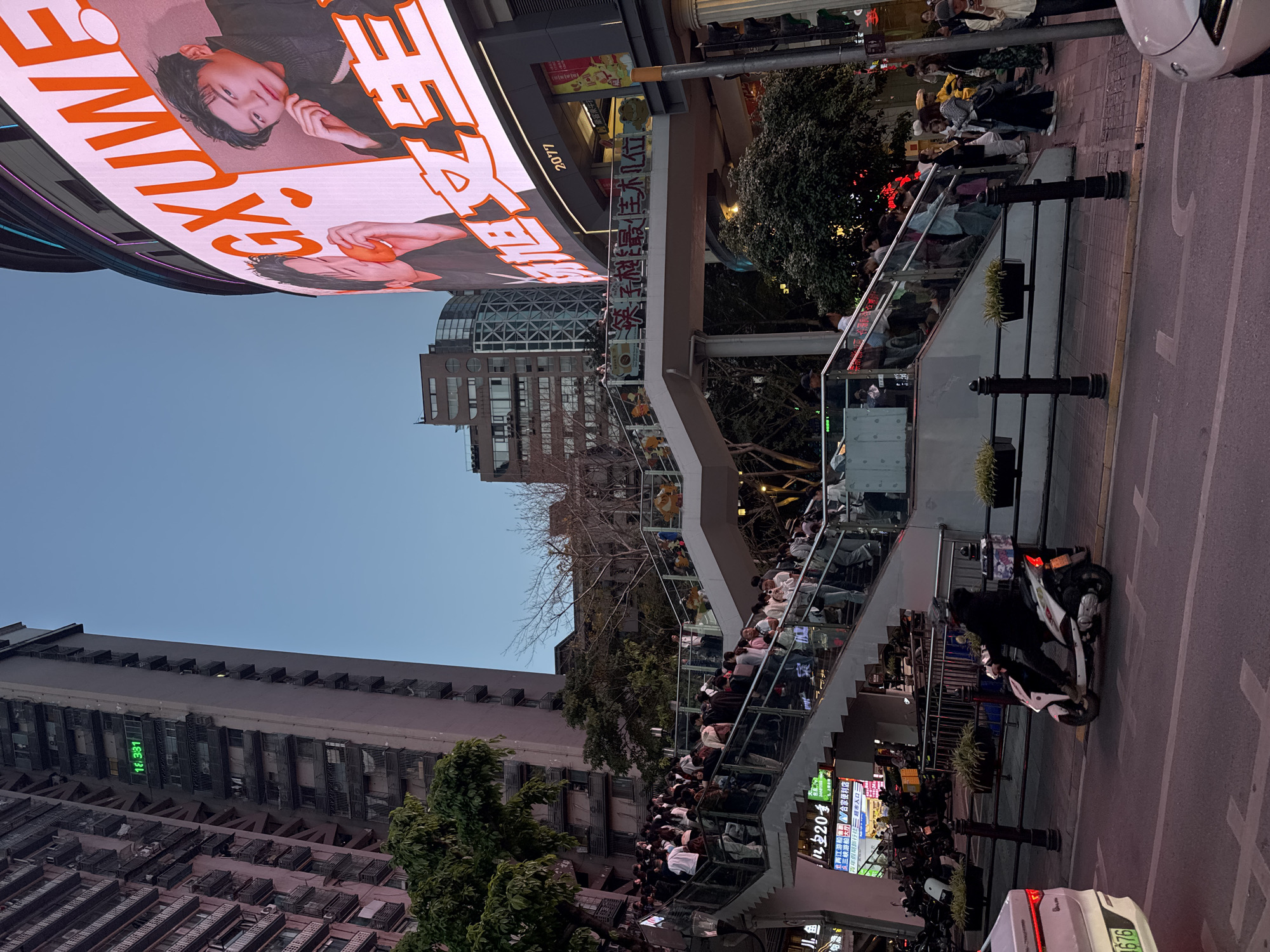 Here is a forensic description of the image:
The image depicts a bustling urban scene in the Yuzhong District of China. A pedestrian overpass is crowded with people. A brightly lit LED billboard displays an advertisement.
Architecture: Several buildings are visible, including high-rise apartments and commercial structures. A prominent building with a digital clock displaying "16:331" is present. The overpass, constructed with glass and metal railings, is heavily populated.
Objects and Subjects: Various motorcycles and a white car are parked along the street. Signs indicating "送子越最佳机位" are visible, alongside advertisements for stores and travel agencies. The billboard showcases the text "GXUWE," with other characters alongside images of a person.
Actions and Interactions: People are seen walking on the overpass. The street appears to be a busy thoroughfare with vehicular traffic.
Environment: The environment is urban, with buildings, trees, and the overpass dominating the landscape. The sky is visible, indicating the image was taken during daylight hours.