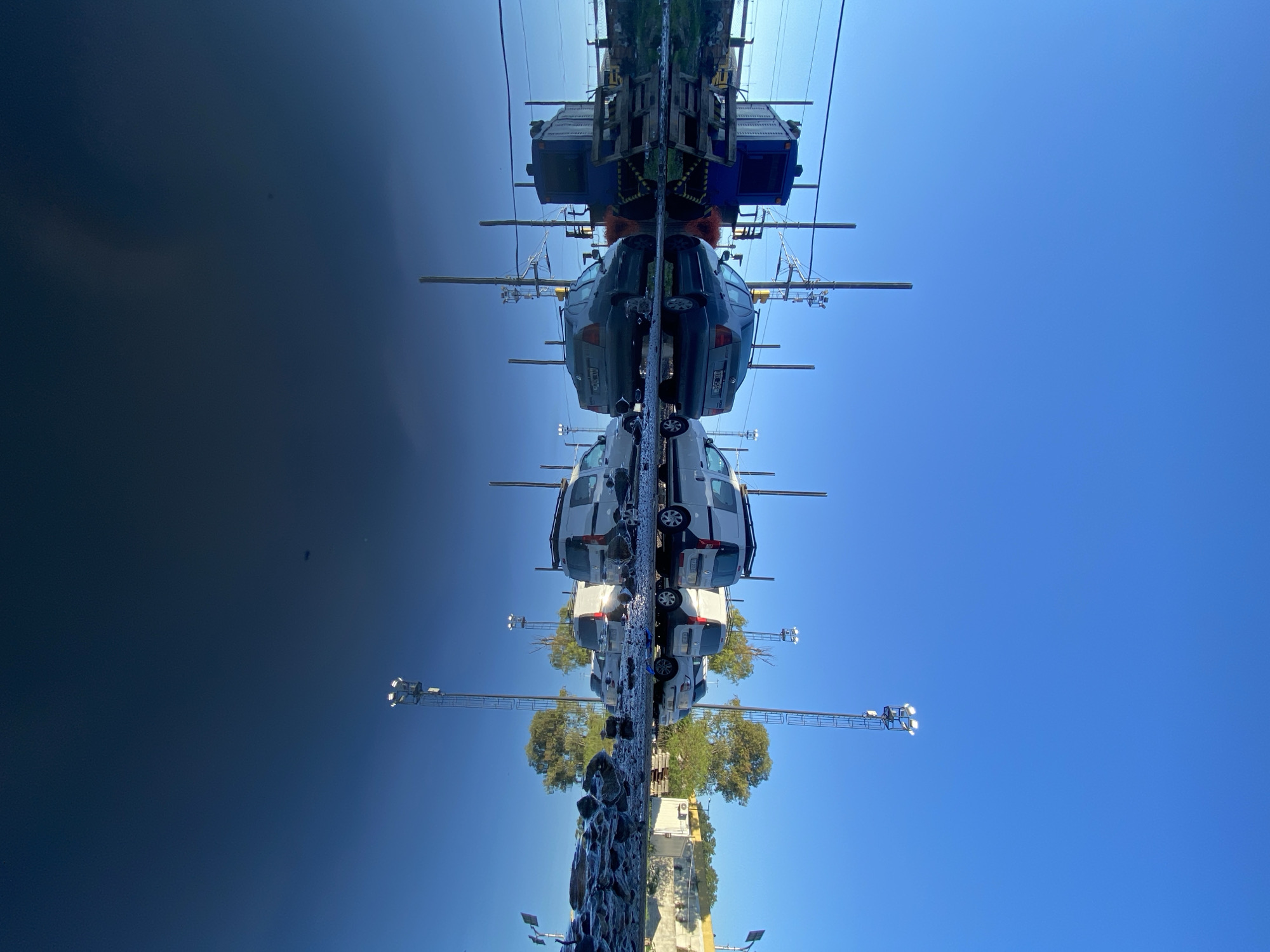 The image shows a reflection of buildings, trees, and cars in the water. The top of the image depicts a bridge with some equipment on top. Beneath the bridge, two rows of cars are visible. Beneath the cars, the image shows a tree, other cars, and some infrastructure. The sky is a clear blue.
The scene is a reflection in water; the city of Buenos Aires, Argentina is not explicitly identifiable. The objects include cars, trees, buildings, infrastructure, and a clear blue sky. There is no evidence of people, actions, or interactions.