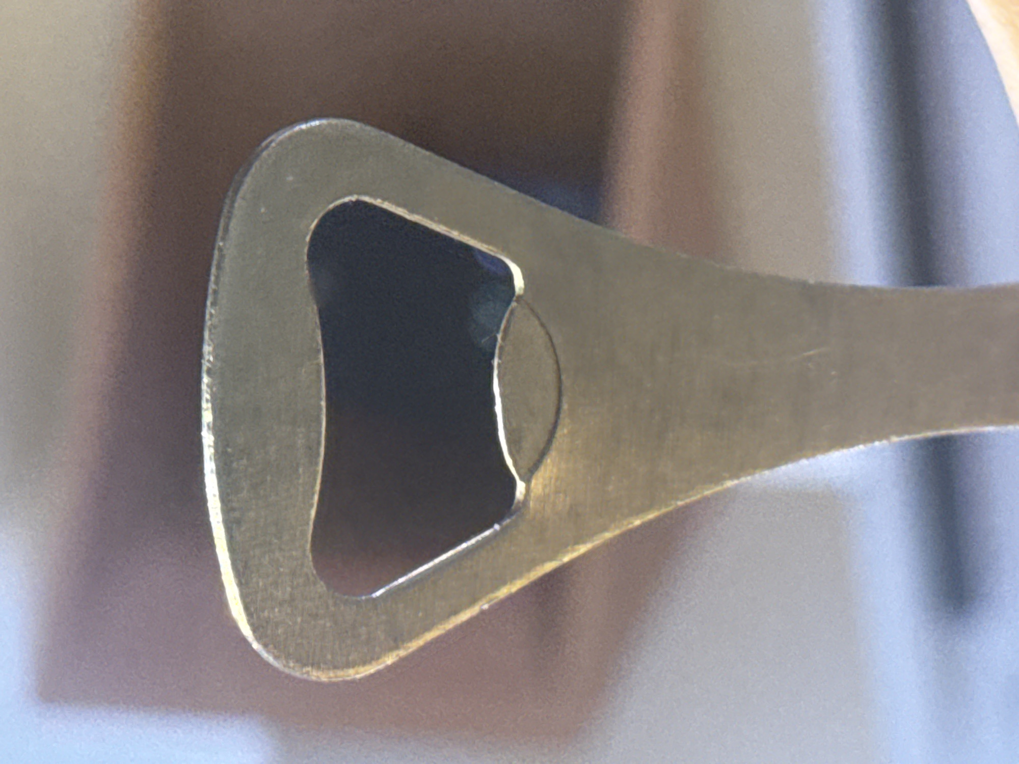 The image presents a close-up, angled view of a metal bottle opener. The object is fabricated from a brass or gold-toned metal, exhibiting a brushed or satin finish with visible surface texture and some minor wear along its edges. The functional end of the bottle opener is the primary subject, featuring the characteristic triangular-shaped opening designed to engage with bottle caps, and a smaller, semi-circular projection within that opening. Light reflects softly off the metal, creating subtle highlights and showing the contours of the object.
The background is completely out of focus, appearing as an abstract blend of soft, neutral colors. These blurry tones consist mainly of gradients of light brown, beige, and hints of a soft blue or gray, suggesting an indoor environment without revealing specific details of the setting. There are no people, activities, or events depicted in the static composition. No specific time of day, weather conditions, or textual elements are visible. The image focuses solely on the form and material of the bottle opener.