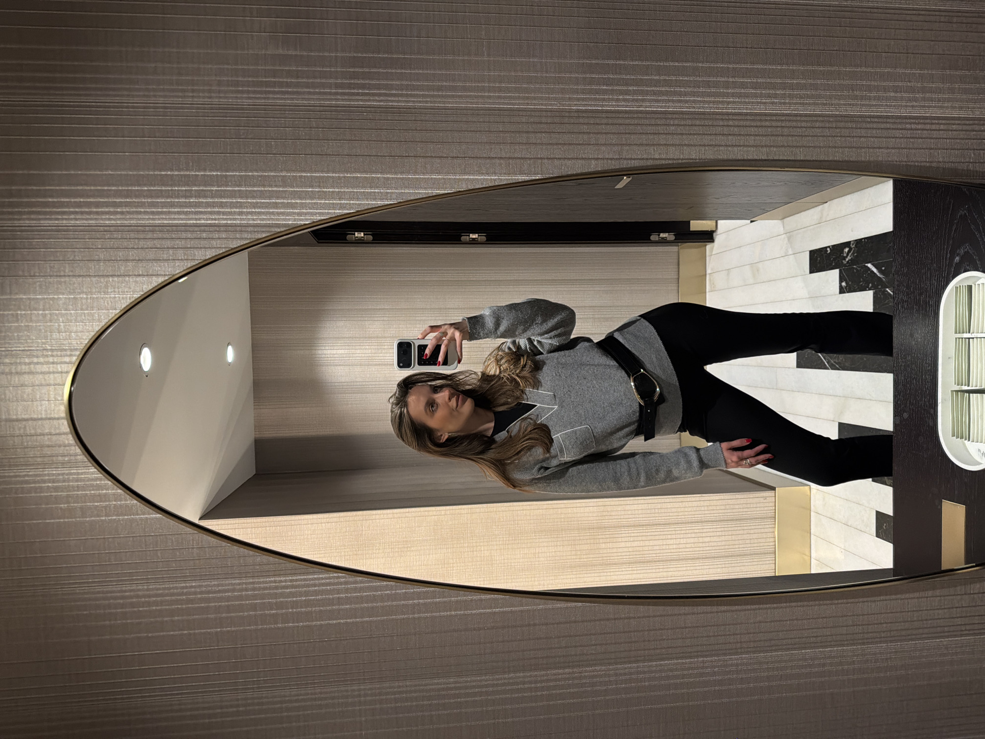 A woman stands in a bathroom, visible in a large oval mirror. She is fair-skinned with long brown hair and wears a gray sweater with a black belt, and black pants. She is holding a smartphone in her right hand, presumably taking a selfie. The bathroom features black and white marble flooring and a textured gray wall. The location is likely in Budva, Montenegro.