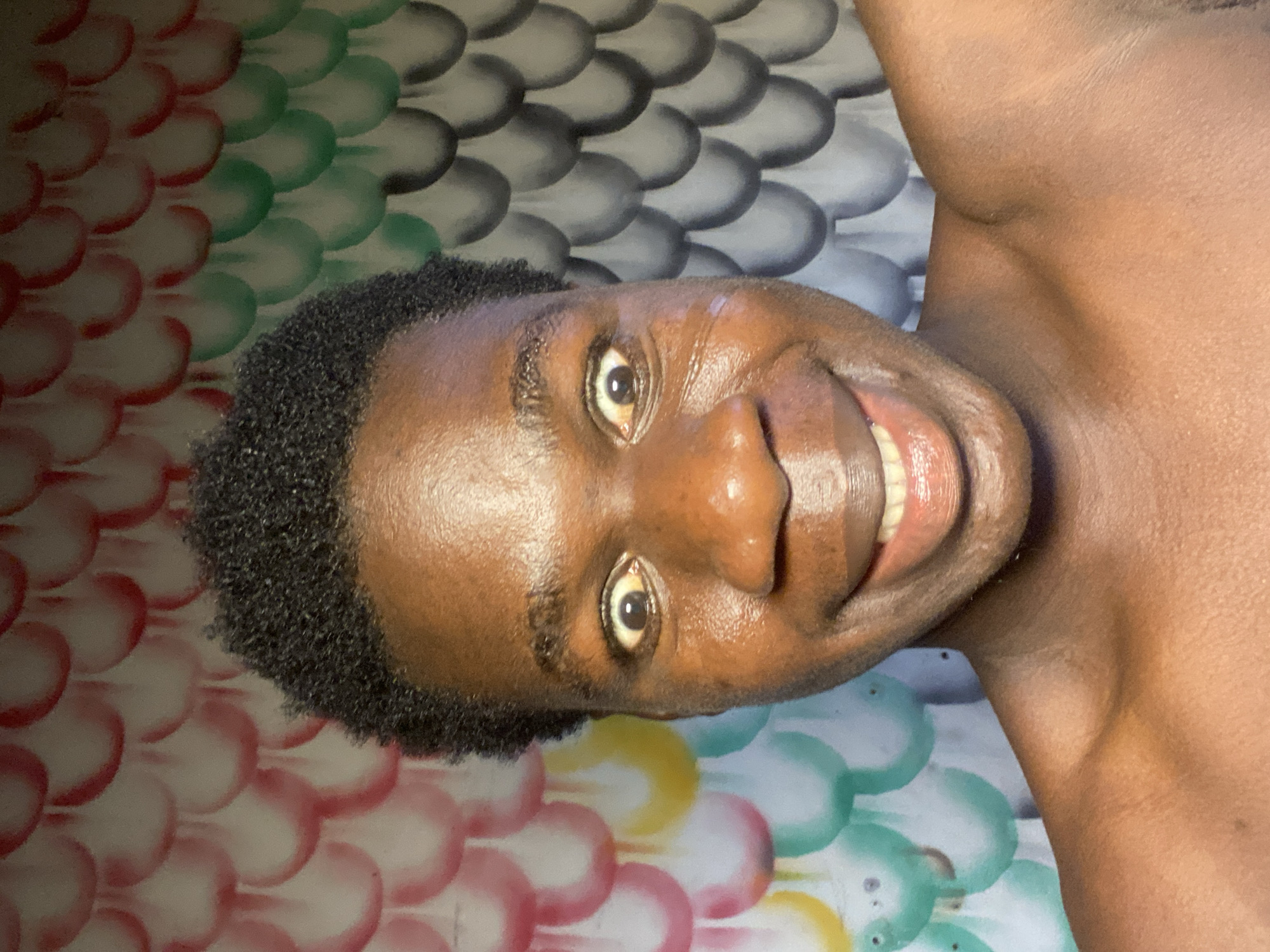 A close-up, upright portrait features a person with dark skin, smiling broadly and looking directly into the camera. The individual, appearing to be an adult, has short, dark, tightly curled hair. A wide smile reveals healthy white teeth, and the eyes are open and bright. The skin on the face and visible upper chest appears smooth and slightly reflective.
The background behind the person is a colorful, patterned surface, likely a wall or fabric. It displays a repetitive design of overlapping, scale-like or scalloped shapes arranged in rows. These shapes are colored in various hues including red, white, green, gray, and yellow. The lighting is bright and even, suggesting an indoor setting.
The subject's expression conveys joy and friendliness. No text is visible in the image. Given the provided context, this scene is set in Mararaba, Nigeria.
