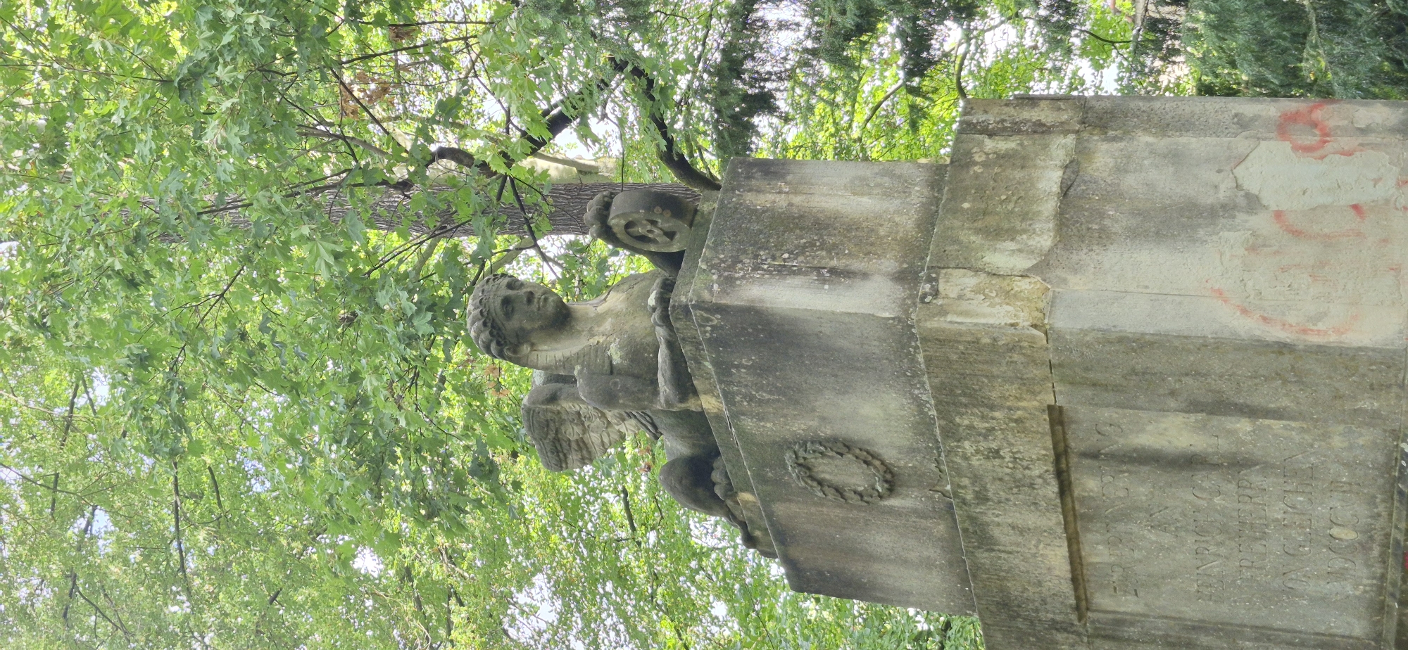 The image shows a close-up view of a weathered stone monument in a park-like setting, with lush green trees forming the background. The monument features a carved stone sphinx with wings, resting on a square pedestal. The sphinx is depicted with a human-like head and the body of a lion. Its face is weathered but discernibly carved. A circular ornament, possibly a laurel wreath, is carved into the pedestal below the sphinx. Further down the pedestal, an inscription is visible, although partially obscured by weathering and damage. The inscription appears to be in German and includes what looks like a date in Roman numerals. The stone of the monument is rough and shows signs of age and exposure to the elements, with patches of discoloration and some red spray paint graffiti visible on the lower section of the pedestal. The overall impression is one of historical significance and natural decay within a verdant environment. Based on available information, this monument is located in the "Vogelherd" park area in Regensburg, Germany, and is believed to be a memorial to Heinrich Carl Freiherr von Gleichen.