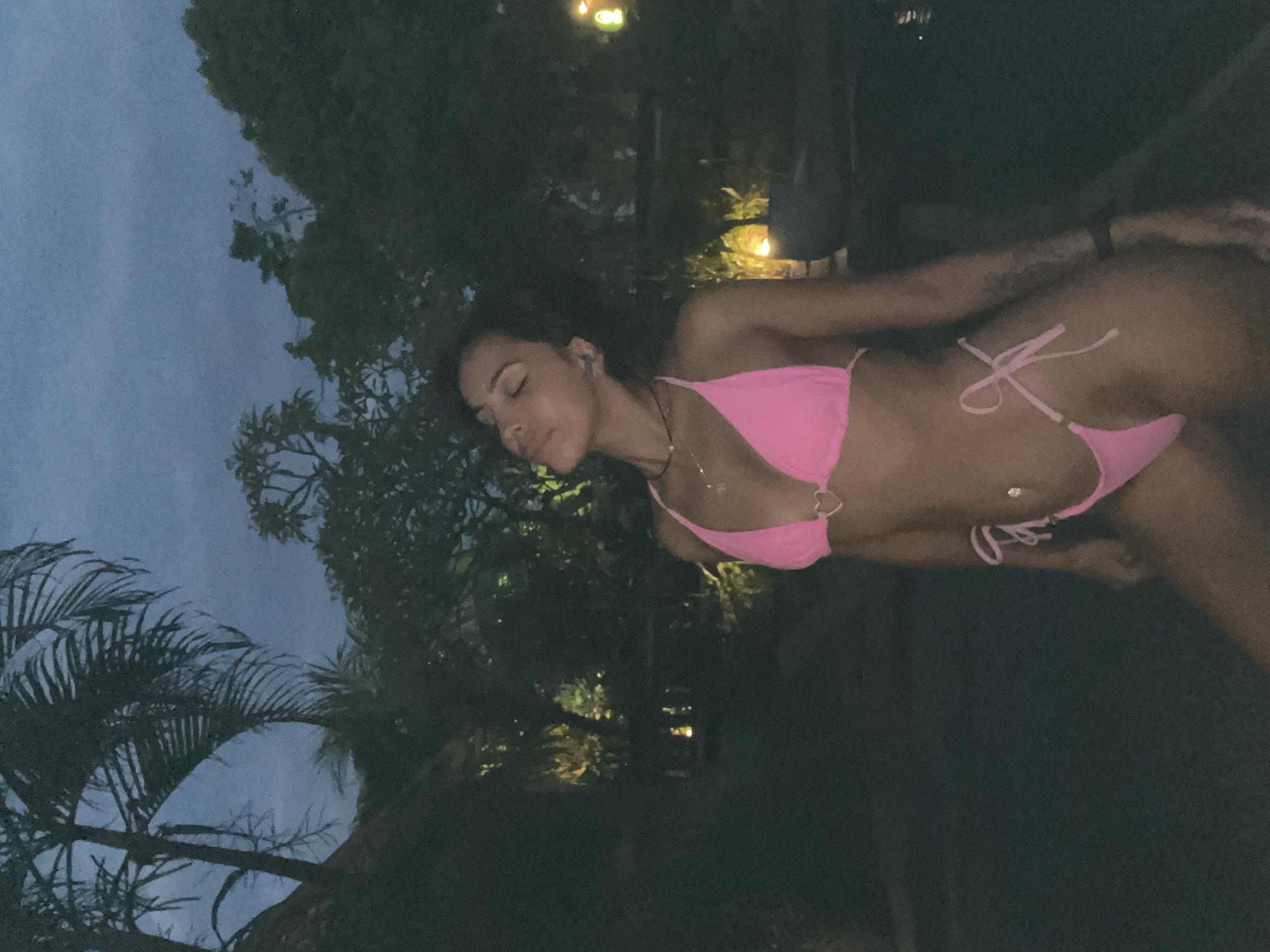 A woman wearing a pink bikini poses in front of vegetation and a dark sky. The location is Kuta Selatan, Indonesia.