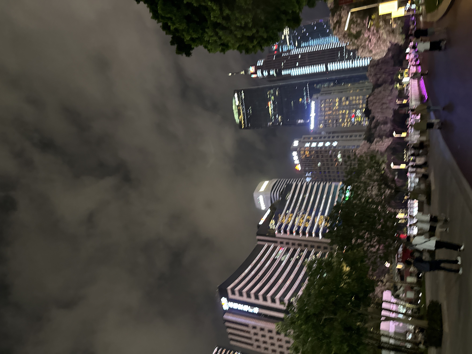 The image depicts a nighttime scene in Tianhe District, China. The foreground features a paved walkway with several people strolling. To the left, trees with sparse foliage are visible, bordering a paved area with lighting. Behind the walkway and trees, a row of modern, illuminated skyscrapers dominates the background. These buildings display various architectural styles and are lit up with a combination of white, yellow, and blue lights. The sky above is dark and cloudy, obscuring any stars or celestial bodies. The overall impression is of a bustling urban environment at night, with a blend of natural elements and towering modern architecture.
