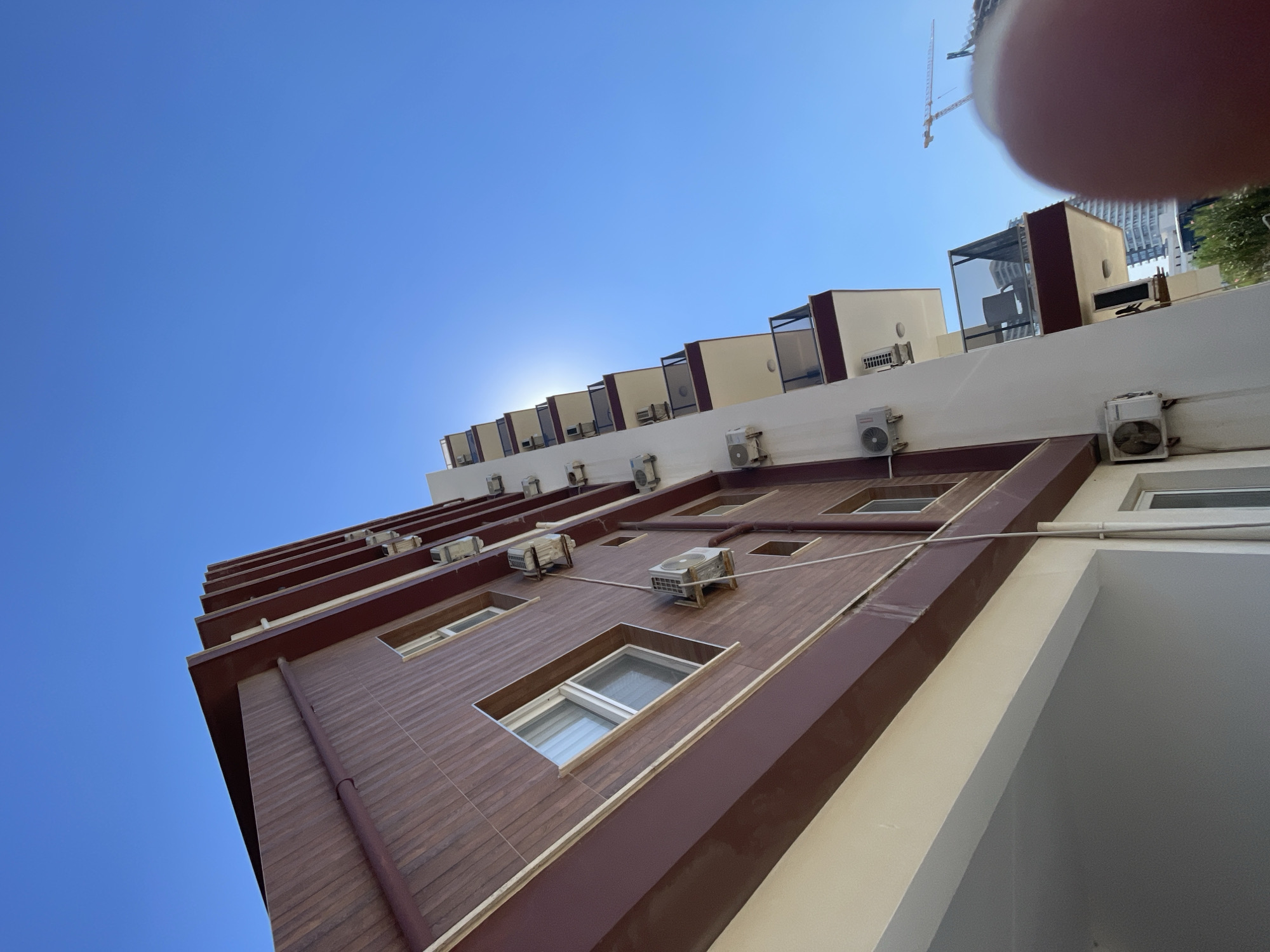 A low-angle, full shot captures the exterior of a multi-story building on a clear, sunny day in Trikomo, Northern Cyprus. The building's facade features a distinctive dark red trim with wood-patterned siding on the upper floors, contrasted with a lighter cream-colored lower section. Numerous white air conditioning units are mounted on the walls of the building, some appearing weathered. Balconies with dark glass railings and red trim punctuate the building's sides. The bright blue sky dominates the upper portion of the image, with the sun partially obscured by the building's structure, creating a lens flare effect. In the background, the skyline includes other buildings and a construction crane, suggesting ongoing development in the area. A blurred fingertip is visible in the lower right corner, partially obscuring the scene.