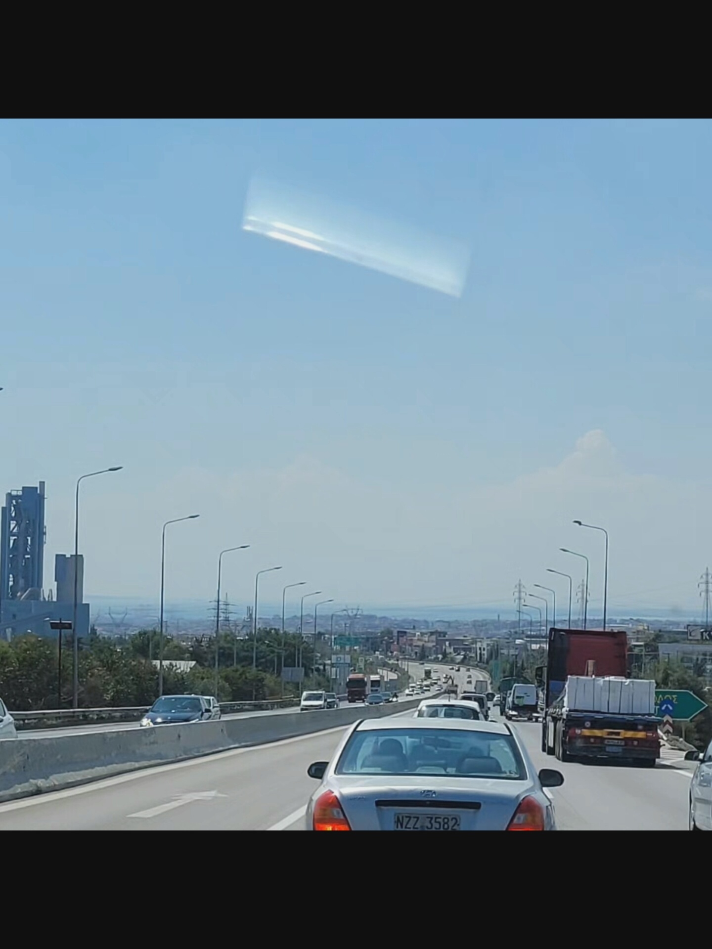 This is a photograph taken from a vehicle moving on a highway, likely during the daytime under a clear, bright sky with some glare. In the upper portion of the sky, there is a prominent, elongated, bright white reflection or anomaly. The highway is multi-lane, with concrete barriers separating directions of travel. Several vehicles are visible on the highway, including cars and trucks. One car in the foreground has a license plate that reads "NZ7 3582". In the distance, a city skyline is visible, featuring industrial structures, buildings, and power lines. The landscape appears somewhat arid with some vegetation. Specifically within the city of Bielawska Wieś, Poland, the scene depicts a section of a highway, characterized by its infrastructure and the flow of traffic.