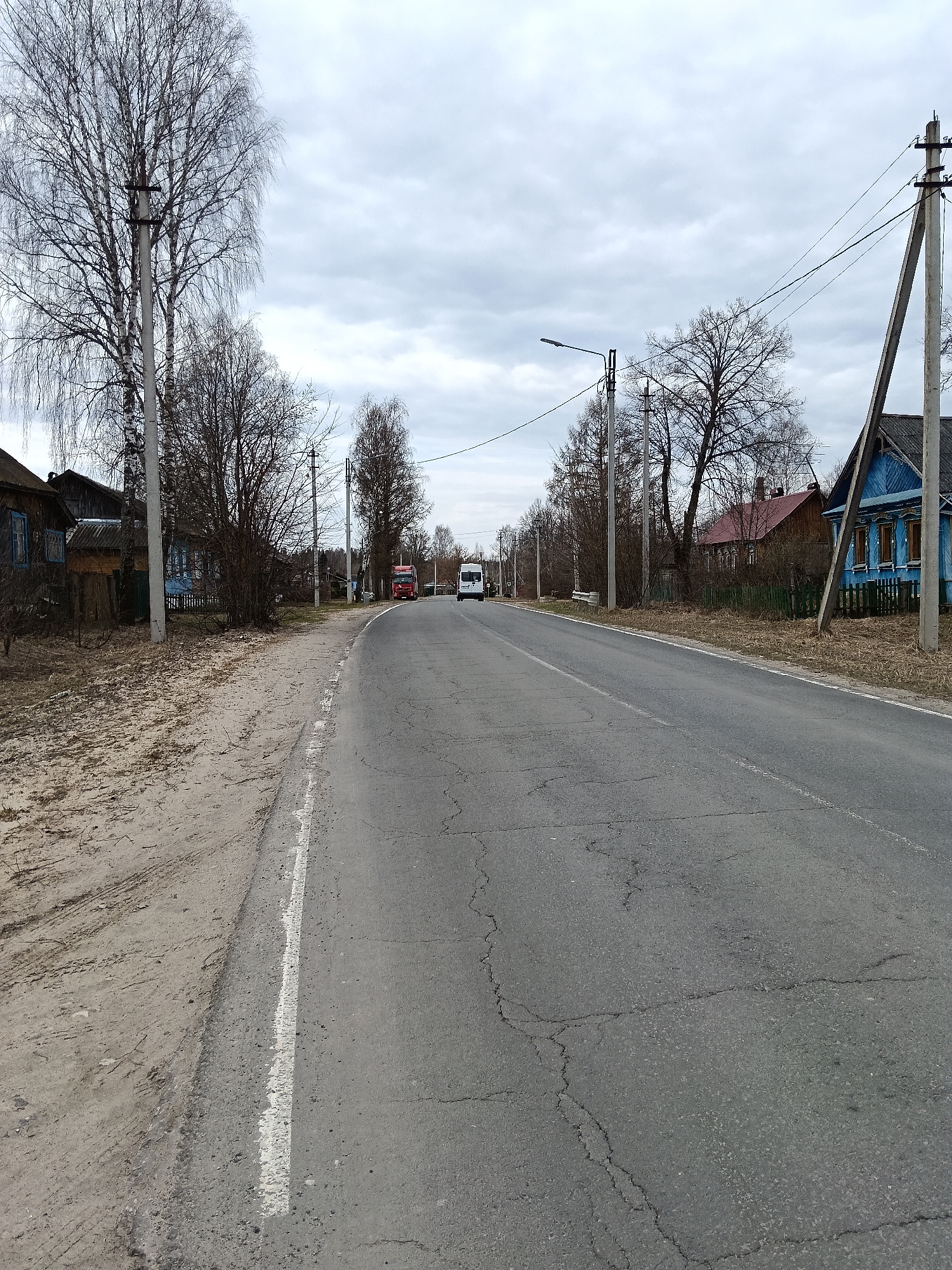 The image shows a straight asphalt road in the village of Красный Маяк, Russia. Cracks mar the road surface. A solid white line marks the road's edge. On either side of the road, bare trees and houses line the street. Utility poles stand along the road, connected by overhead wires. In the distance, a white van and a red truck are visible on the road. The sky is overcast.