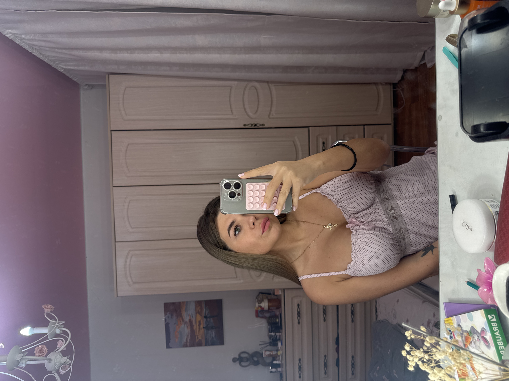 A female takes a mirror selfie in a room. The room has light pink walls and white furniture including a wardrobe and dresser. A chandelier is visible in the lower left. A painting hangs on the wall behind the dresser. The location is implied by the inclusion of the Russian-language textbook on the right, and is Chaikovsky, Russia.