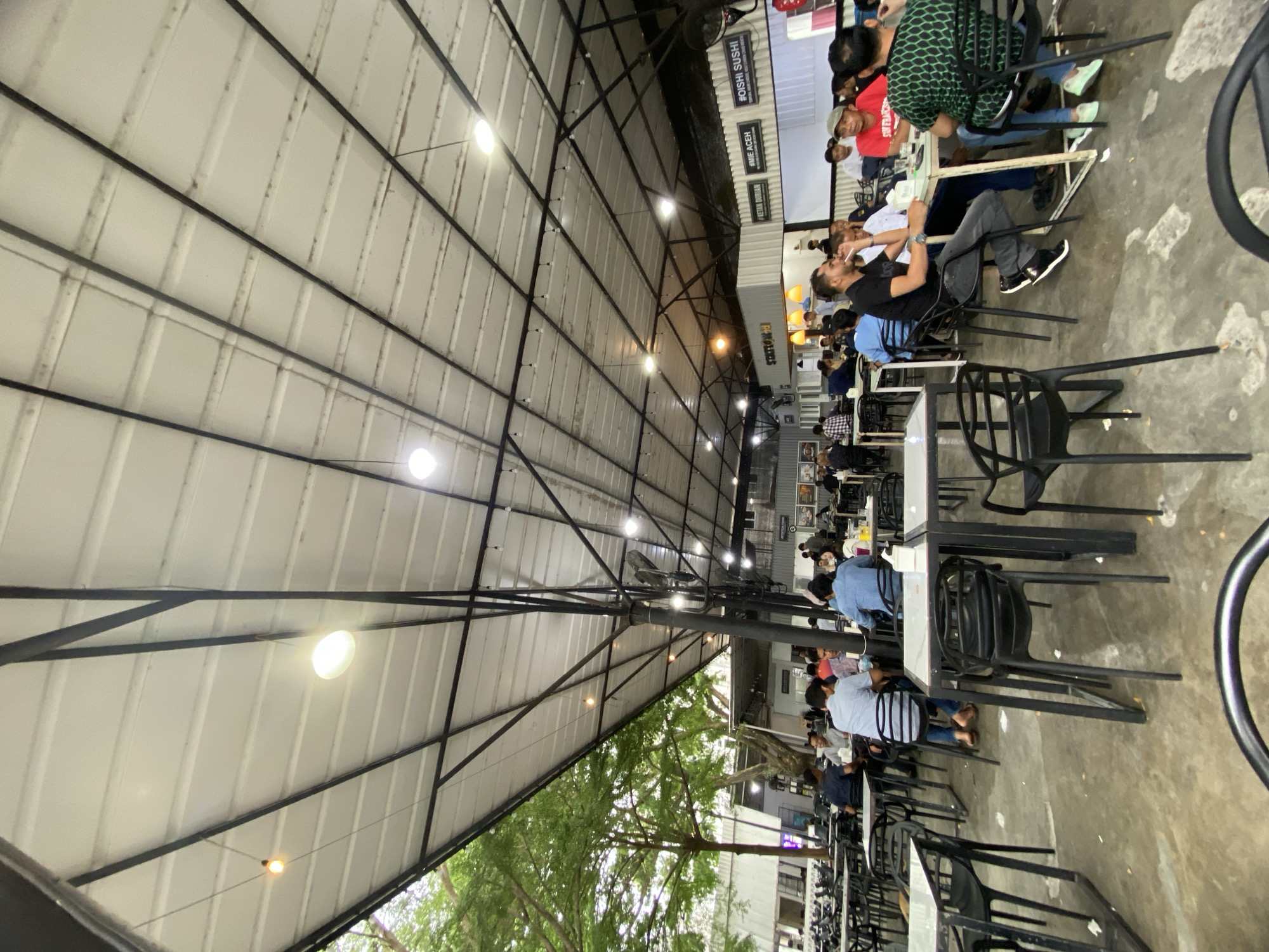 The image captures a bustling semi-outdoor dining area, likely a food court or a collection of small restaurants, under a shared canopy in Lhokseumawe, Indonesia. The scene is illuminated by numerous artificial lights, indicating it is evening or night.
Many people, mostly adults, are seated at rectangular tables with black chairs, engaged in dining, socializing, and relaxing. Patrons are dressed in casual clothing. In the midground, a man in a black t-shirt and dark pants is prominently seated, holding an object, while other diners are visible in various states of interaction throughout the space. The atmosphere appears lively and social.
The entire area is covered by a large, white, corrugated metal roof supported by a black metal truss framework. Multiple bright, circular LED lights are affixed to this structure, casting strong illumination across the scene. Additionally, smaller, warm-toned string lights are visible. The ground is a light-colored concrete or paved surface. On the left side of the frame, green foliage from trees can be seen, emphasizing the open-air aspect of the establishment.
Along the far wall on the right, several signs are displayed, advertising food options. Visible text includes "#OISHI SUSHI," "#MIE ACEH," and "#PRETAM DURIAN." The inclusion of "Mie Aceh," a popular dish from the Aceh region of Indonesia, strongly supports the given location context of Lhokseumawe. The varied signs suggest a diverse range of culinary choices available to customers at this popular gathering spot.