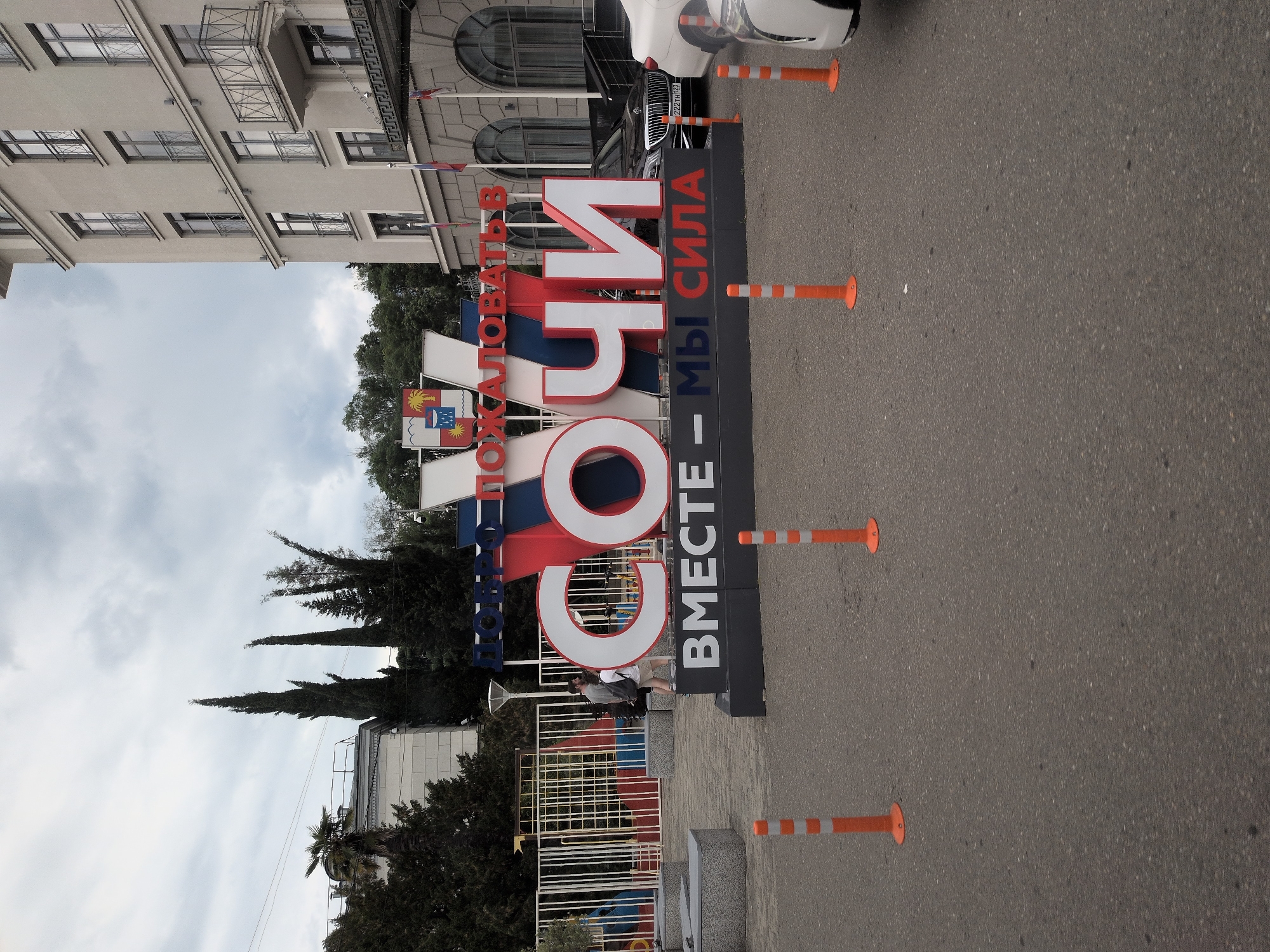 This image depicts a large, prominent sign in the city of Sochi, Russia, that reads "ДОБРО ПОЖАЛОВАТЬ В СОЧИ" (Welcome to Sochi) with the additional phrase "ВМЕСТЕ - МЫ СИЛА" (Together - We are Strength) below it. The sign is made of large, three-dimensional letters, with "СОЧИ" being the most visually dominant. The letters are in white and red, with blue accents at the top of the sign. The sign is situated outdoors on an asphalt surface, with several orange traffic cones placed around it.
Behind the sign, a multi-story building with a light-colored facade and numerous windows and balconies is visible. Flags are mounted on the building's exterior. The sky is overcast and cloudy. In the background, there are trees, including tall cypress trees and a palm tree. A black car, possibly a Mercedes-Benz, is parked to the right of the sign, with a partial view of a white car to the far right. Two people, a man and a woman, are walking near the sign. The overall scene appears to be an outdoor public space, likely a tourist area or an entrance to a facility in Sochi.