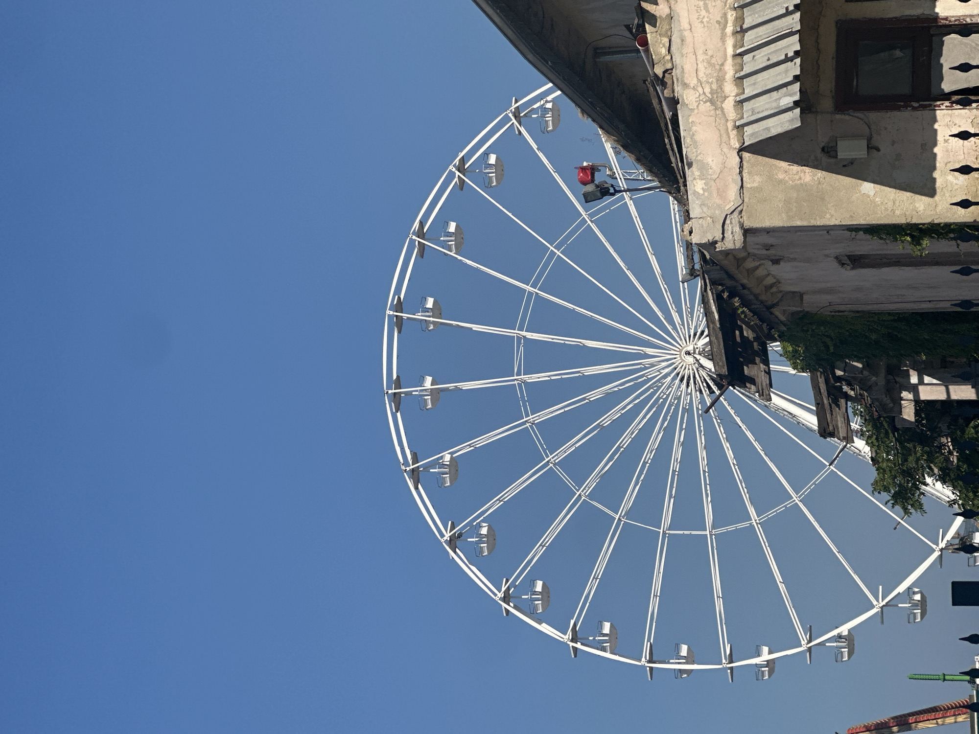 A white Ferris wheel is visible against a clear blue sky. The wheel is large and circular, with many spokes and gondolas attached. In the foreground, there is a partially visible dilapidated building with a weathered facade and a corrugated metal roof. Green vines grow on the side of the building. The scene appears to be outdoors in a bright, sunny environment.