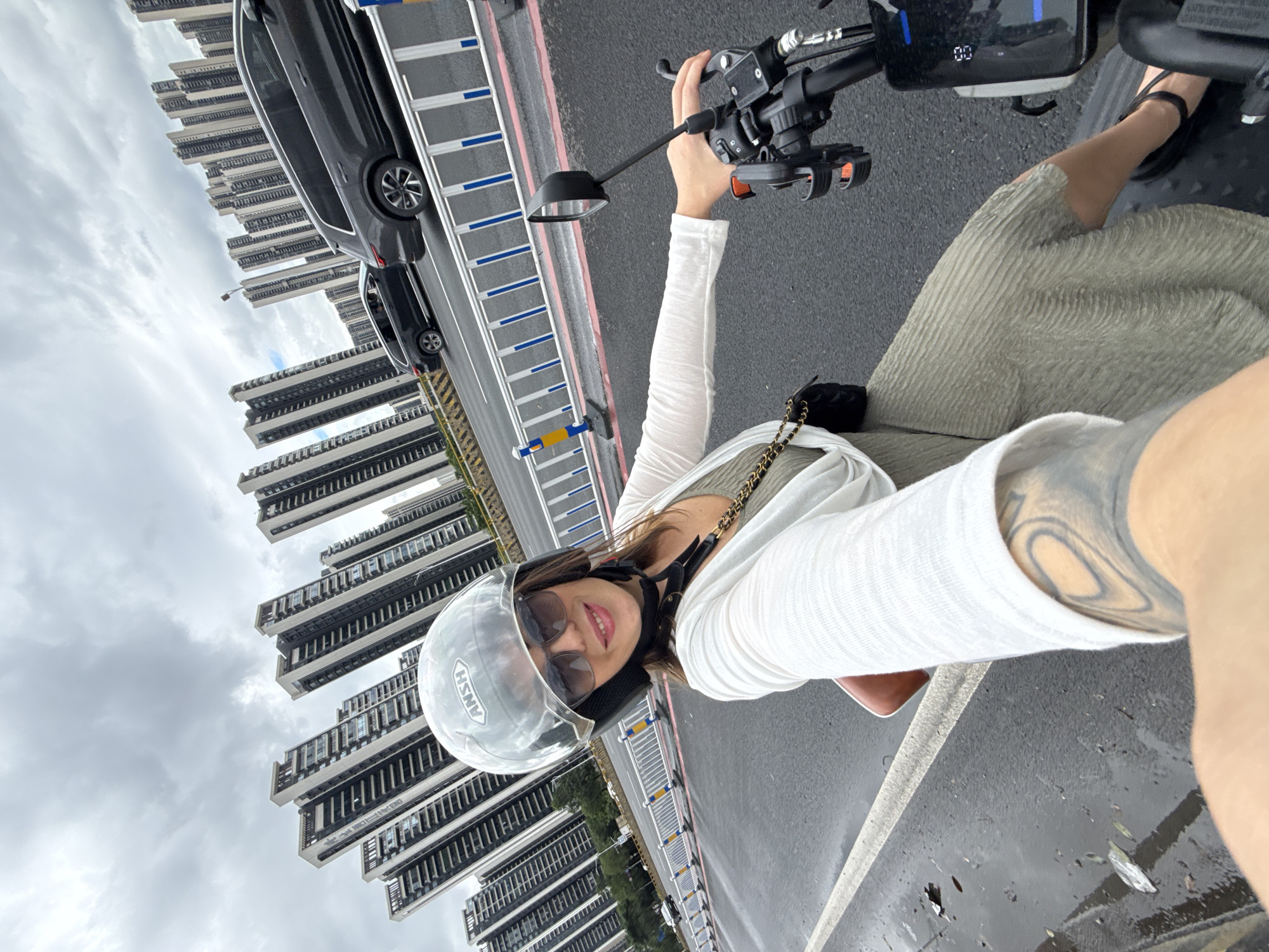 In Huizhou, China, a woman wearing a helmet and sunglasses is riding an electric scooter on a wet road. She is taking a selfie, with the scooter's handlebars and a portion of the vehicle's display visible in the foreground. The woman is wearing a white cardigan over a green dress and has tattoos on her left arm. The background is dominated by a row of tall, modern apartment buildings under a cloudy sky. Cars are visible on the adjacent road, separated by a white barrier with blue accents.