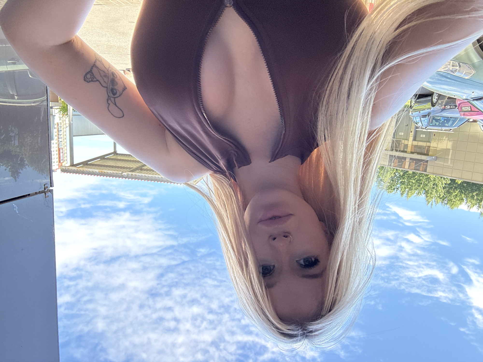 A young woman with blonde hair and a nose ring is taking a selfie outdoors. She is wearing a brown, sleeveless top with a zipper that is partially undone, revealing her chest. A tattoo of a cartoon character is visible on her upper left arm. In the background, there are cars parked in front of a building with a light-colored facade. Trees and a blue sky with scattered clouds are also visible. The overall scene suggests an urban or suburban environment on a sunny day. The location is not specified beyond being in the city of Saint Petersburg, Russia.