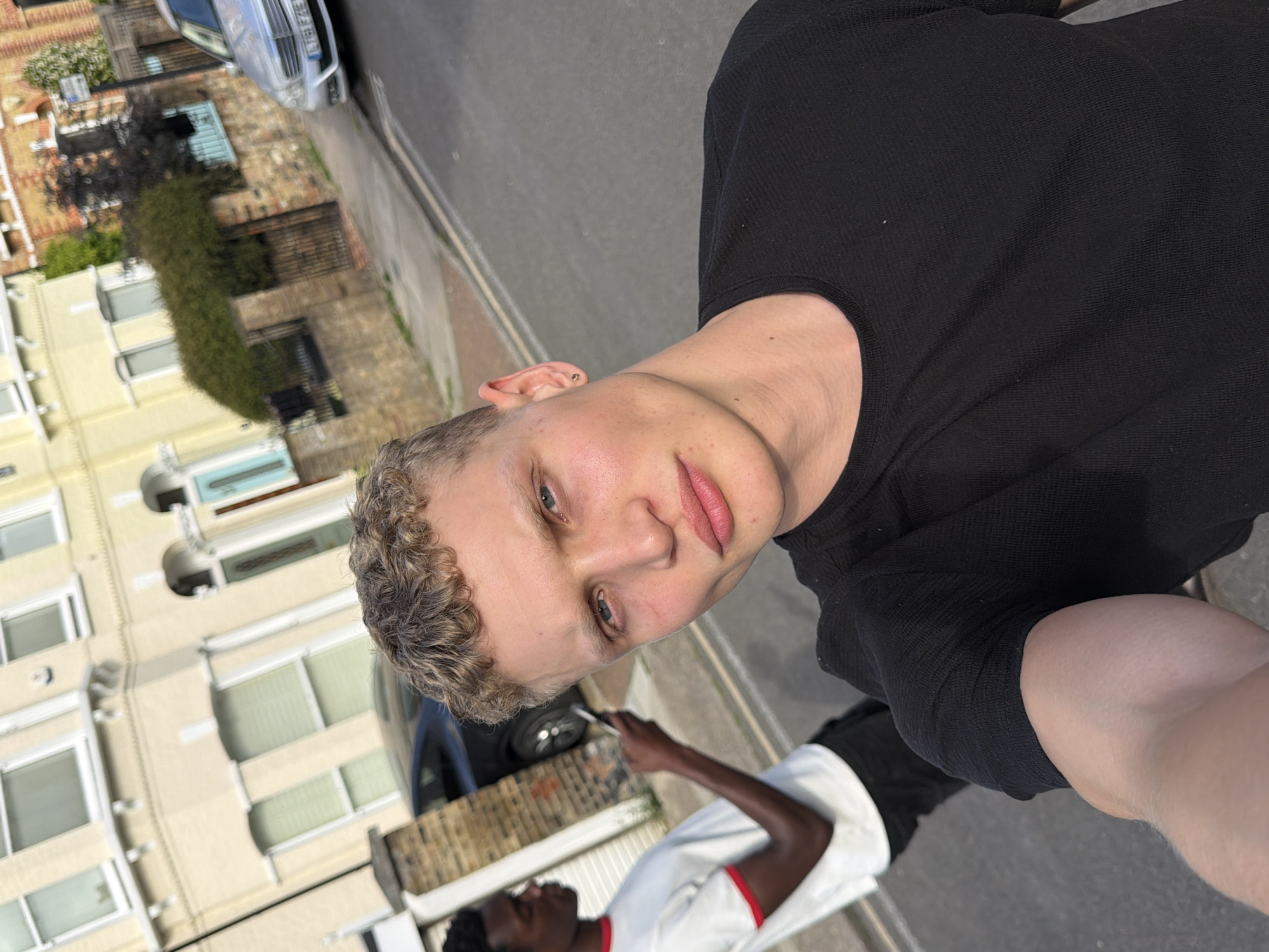 Two young men are standing on a residential street in London, United Kingdom. The man in the foreground is facing the camera and taking a selfie. He has short, curly blonde hair and is wearing a black t-shirt. He has blue eyes and full lips. The man in the background is facing away from the camera, looking at his phone. He is wearing a white t-shirt with red trim. The street is lined with terraced houses, some of which are painted yellow. A silver car is parked on the street in the background. The sun is shining, and the lighting suggests it is late afternoon. The overall scene depicts a casual, everyday moment.