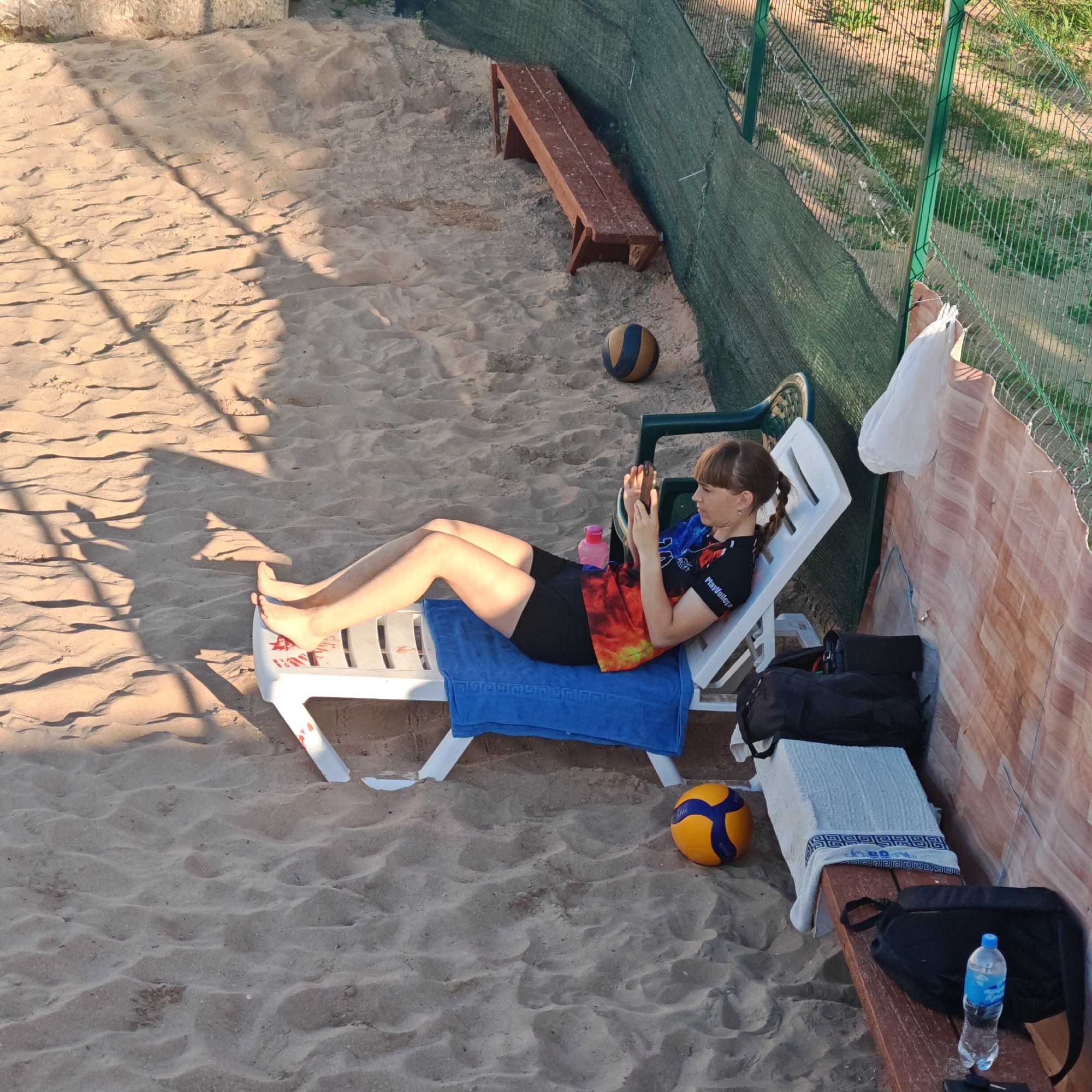 A woman with braided brown hair, wearing a black t-shirt with a tie-dye design in orange and yellow and black shorts, is lying on a white reclining beach chair covered with a blue towel. She is holding a mobile phone in her hands and looking at it. The beach chair is on a sandy surface, which appears to be a beach volleyball court, as evidenced by the presence of a volleyball near the chair and another one further away next to a wooden bench. The background features a green fence and some vegetation. The scene appears to be outdoors, likely during the daytime due to the shadows cast by the beach chair. The location is specified as Gorodskoye Poseleniye Shchelkino, Ukraine.