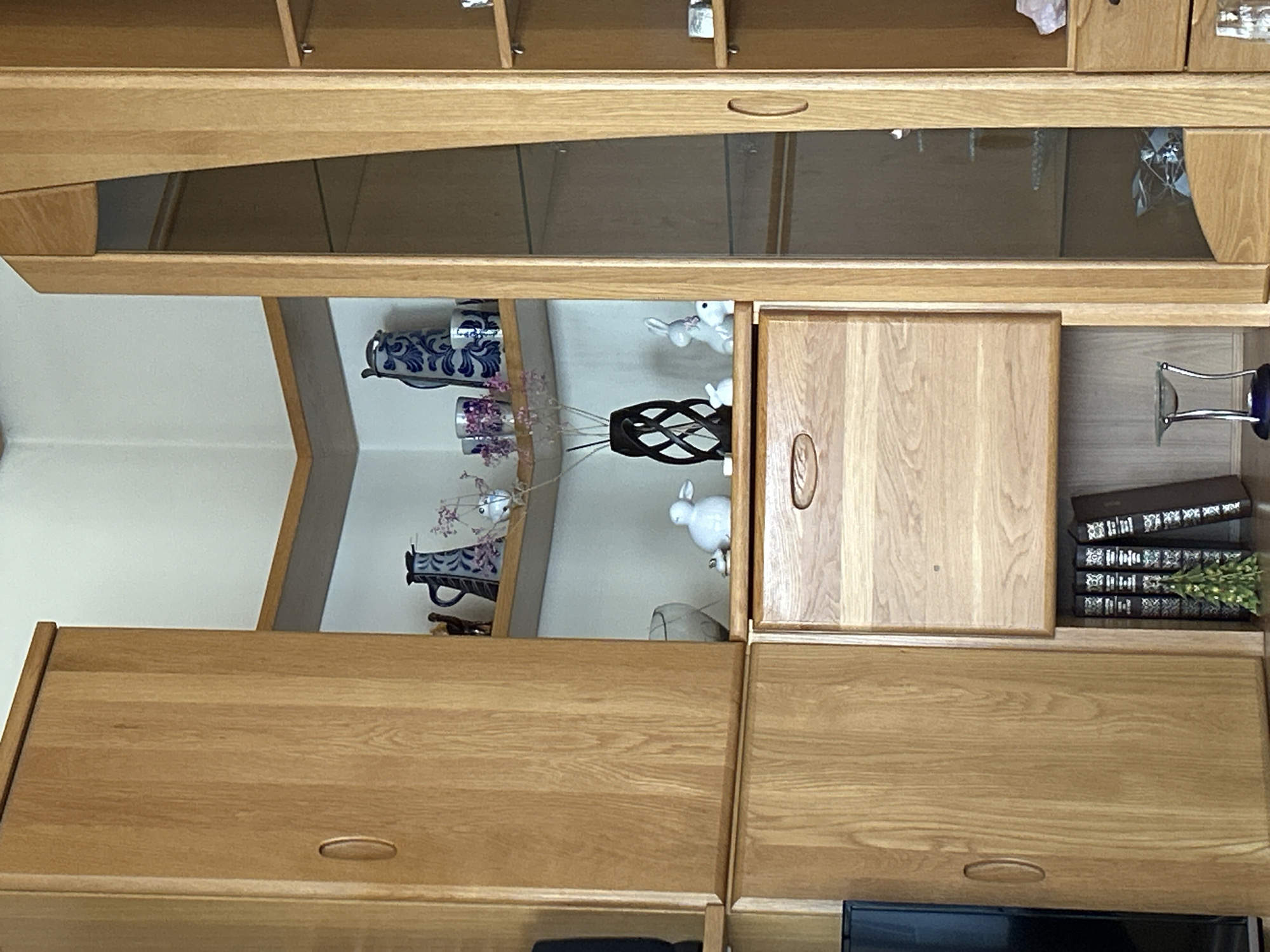 The image shows a section of a wooden furniture unit, likely a cabinet or shelving unit, made of light-colored wood. The unit is positioned in an interior space with a light-colored wall.
On the top shelf of the unit, there are several decorative items, including a blue and white patterned pitcher, two small clear glasses with purple contents, and a small pink flower arrangement. To the right of these, on a higher shelf, are two more small decorative items.
Below this top shelf, on a slightly lower shelf, are a white ceramic rabbit, a smaller white ceramic rabbit, and a black decorative vase with an intricate spiral design. There is also a clear glass bowl visible.
Underneath this shelf, there is a wooden drawer with a handle. Below the drawer, a collection of books is visible, with spines facing outward. A small green decorative Christmas tree is placed in front of the books. To the far right of this section, there is a glass-fronted cabinet with shelves. On these shelves, some indistinct items are visible, including what appears to be a pink crystal and some glass objects.
The lighting in the image suggests an indoor environment, possibly during the day. The overall impression is one of a domestic interior with displayed decorative items.