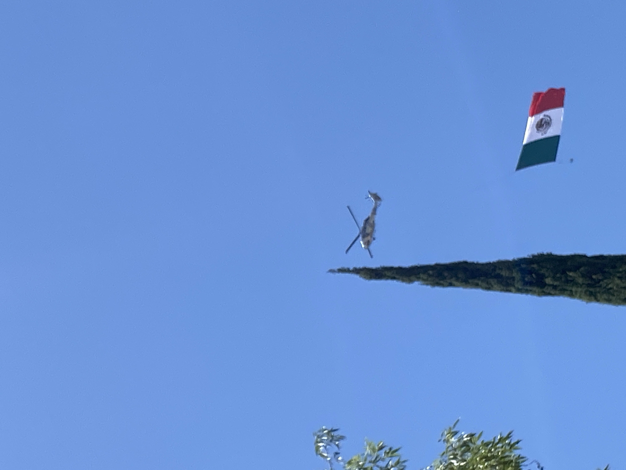The image captures an outdoor scene under a clear, bright blue sky, likely in Puebla, Mexico, during daylight hours.
The primary subjects are a helicopter and a large Mexican flag, both in motion against the sky. The helicopter, predominantly silver or light grey, is seen from below, angled sharply as if banking or in a dynamic maneuver. Its main rotor blades are visible, and the tail boom extends upwards.
To the right of the helicopter, a substantial Mexican flag is unfurled and clearly visible. It features the characteristic vertical stripes of green, white, and red, with the national emblem—an eagle devouring a snake on a prickly pear cactus—prominently displayed in the white central stripe. The flag appears to be pulled or towed, suggesting it's part of an aerial display, possibly for a ceremonial event or parade.
In the mid-ground, a dense, dark green treeline runs horizontally, partially obscuring the helicopter. Another patch of lighter green foliage is visible in the bottom right corner, indicating more trees below the frame. The overall impression is one of a public aerial display or flyover, characterized by the prominent national flag. There is no visible text in the image.