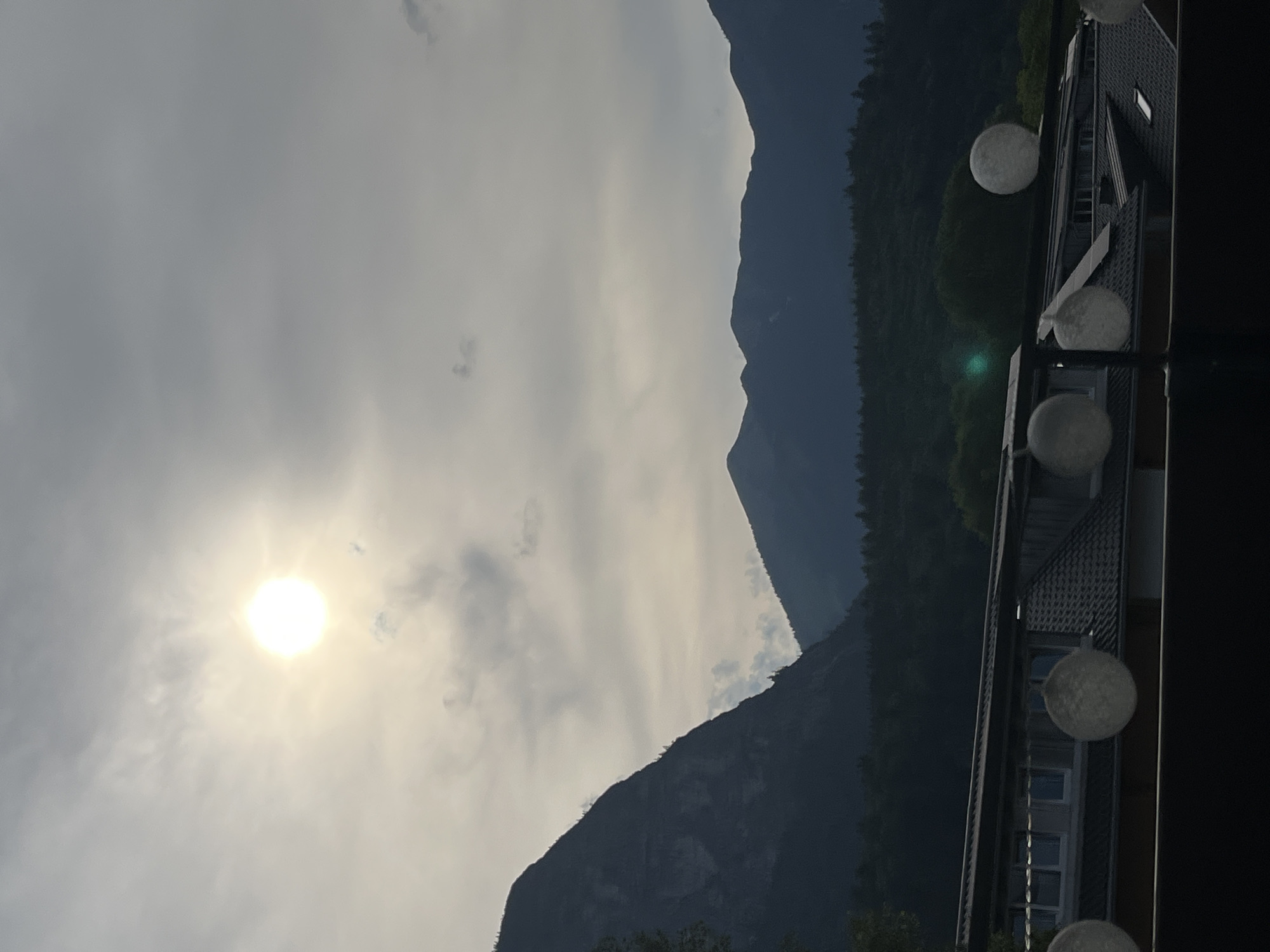 This media file depicts a mountain landscape with a sunlit sky. The main subjects are majestic mountains, partially obscured by clouds, and the bright sun. In the foreground, to the right, a modern building with a dark roof and several windows is visible, along with decorative spherical lights. The scene is likely a scenic overlook or a residential area with a view of the mountains.
The time of day appears to be late afternoon or early evening, given the soft, diffused sunlight and the atmospheric haze. The weather is partly cloudy, with the sun shining through thin clouds, creating a bright, slightly hazy effect. The location context provided, Garmisch-Partenkirchen, Germany, suggests the Alps as the mountain range, known for their dramatic peaks and picturesque scenery.
There are no people or specific activities evident in the image. The notable details include the stark contrast between the bright sky and the dark, imposing mountains, and the clean lines of the modern building. The spherical lights add a touch of contemporary design to the natural backdrop. The overall mood is one of serene natural beauty, with a hint of human presence through the architecture. No visible text is present in the image.