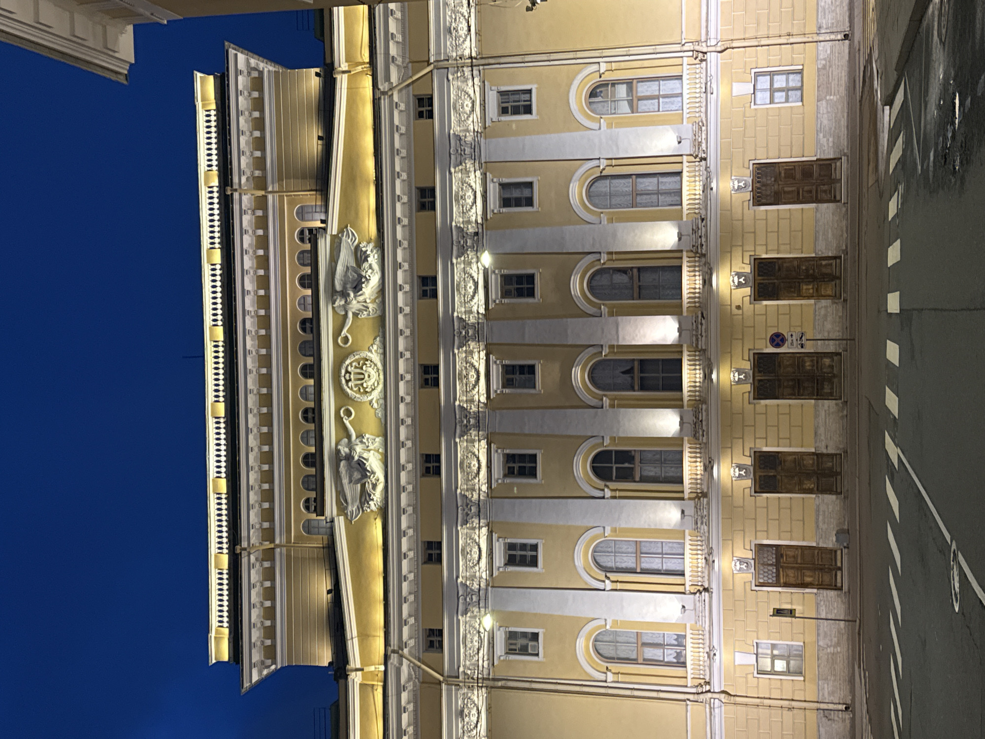 The image depicts the facade of a classical-style building in Saint Petersburg, Russia, likely illuminated at night. The structure is characterized by a yellow exterior with white architectural details, including arched windows and decorative moldings. Sculptural elements such as figures or emblems are present on the facade. A street with crosswalk markings is visible to the right of the building. The sky in the background is a dark blue.
