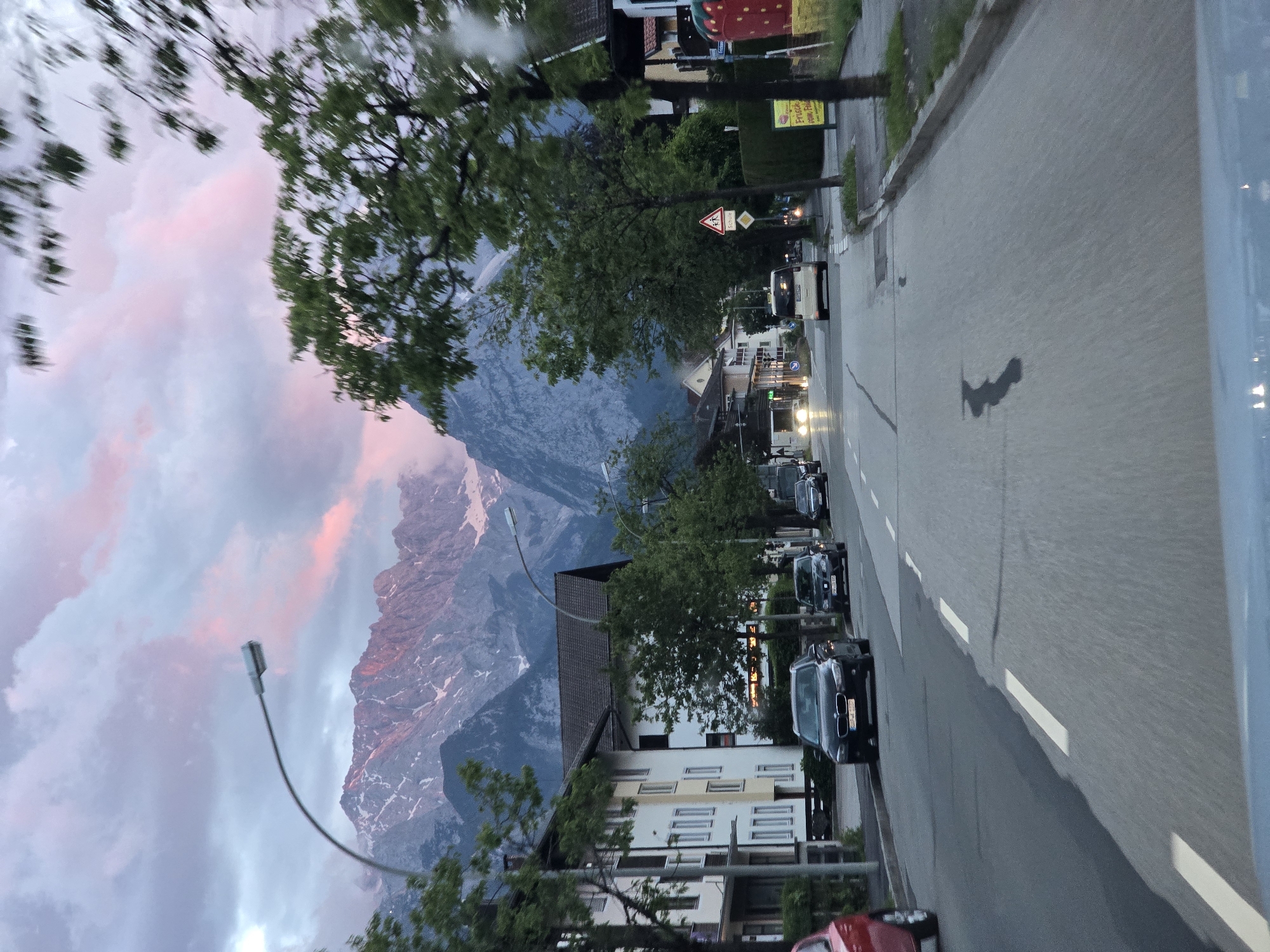 The image captures a street scene in Garmisch-Partenkirchen, Germany, with the Zugspitze mountains in the background. A road runs through the center of the frame, with cars parked and driving on either side. Buildings line the street, some appearing to be residential and others commercial. Trees are present along the roadside, some casting shadows on the pavement. The sky is overcast with streaks of pink and purple, suggesting either sunrise or sunset. A street lamp is visible on the left side of the image, and a traffic sign indicating a school crossing is present on the right. The overall impression is that of a quiet, residential street in a mountainous region.