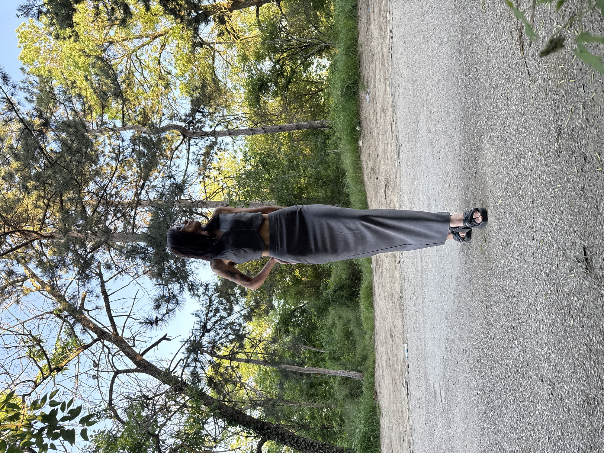 A woman stands on an asphalt road in a park-like setting. She is wearing a dark gray two-piece outfit consisting of a crop top and a long skirt, along with black sandals. The surrounding environment includes trees, foliage, and blue sky visible through the tree canopy. The location is identified as being in Pleven, Bulgaria.