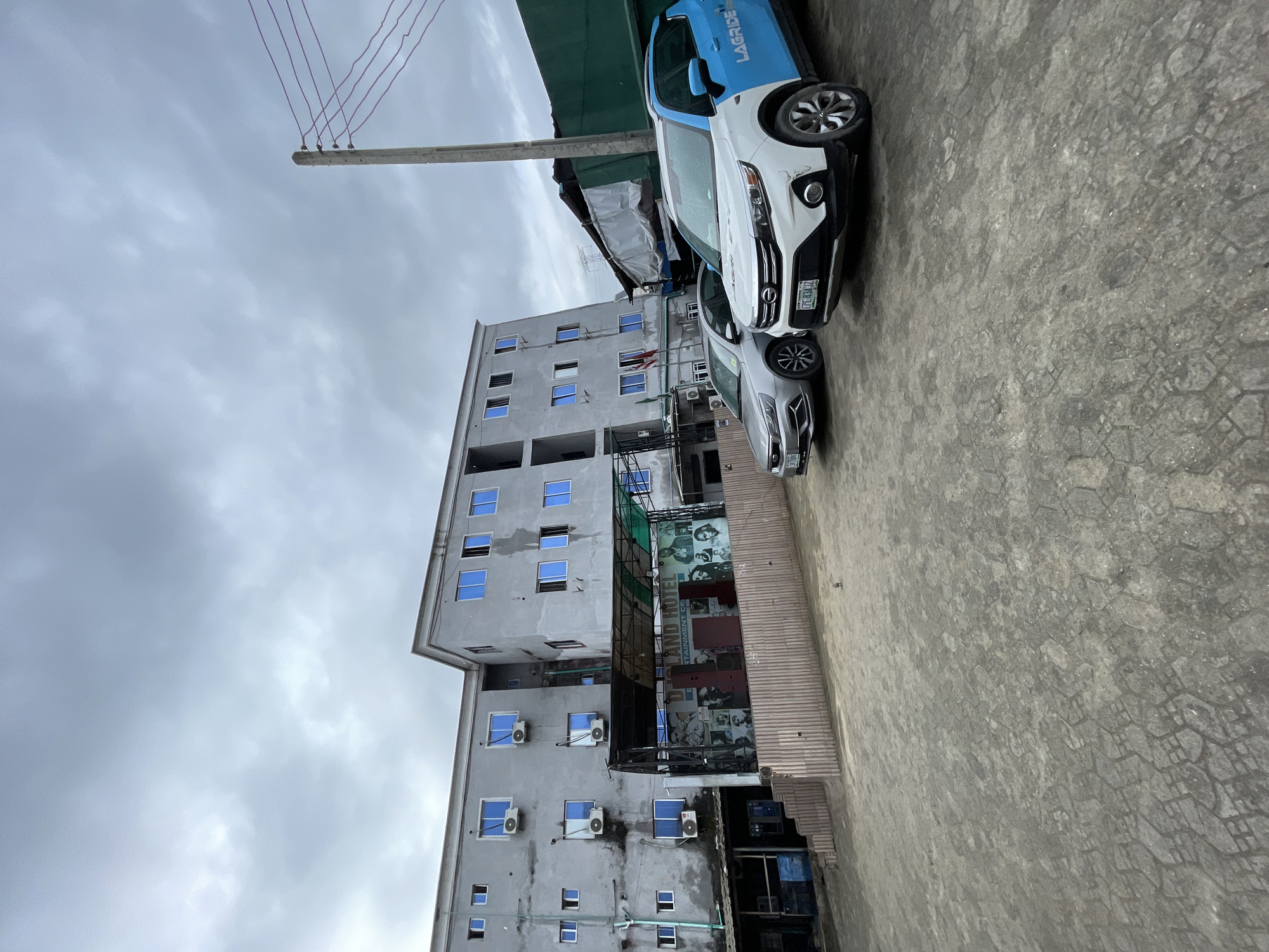 This is a slightly overcast, eye-level, outdoor, full-shot photograph of the entrance to the "DJOLAND HOTEL" in Ado, Nigeria. The hotel is a multi-story building with a light gray facade and blue-tinted windows. Numerous air conditioning units are mounted on the exterior walls. In front of the hotel, there is a covered outdoor stage area with a dark roof supported by metal poles. The stage backdrop features a collage of images, including portraits of various individuals and text that appears to include "ENTERTAINMENT CE". Two cars are parked in front of the stage area: a silver sedan on the left and a white and blue vehicle with "LAGRIDE" branding on the right. The ground in the foreground is paved with hexagonal interlocking stones. A concrete utility pole with multiple wires runs along the right side of the frame, and other structures, including a green corrugated metal wall, are visible behind the cars. The sky is cloudy, and the overall lighting is diffused.