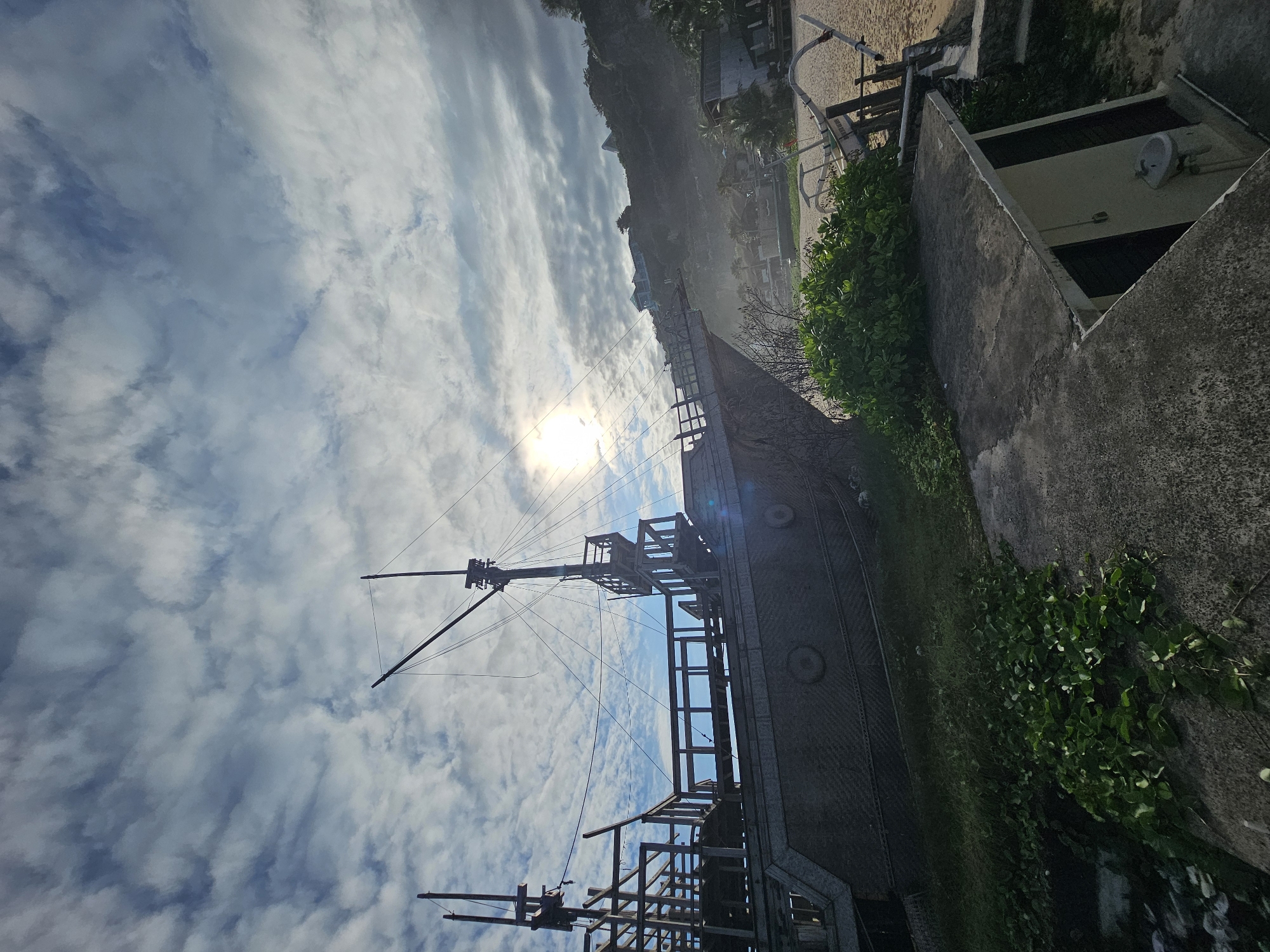 The image shows a ship with masts extending towards the sky. The sky is blue with white clouds and the sun is visible. Vegetation grows on a concrete wall next to the ship. In the background are buildings and a sandy beach. The location is Kuta Selatan, Indonesia.