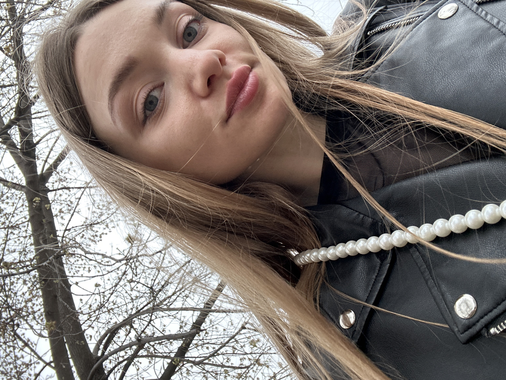 A young woman with long blonde hair, fair skin, and light eyes is posing in a selfie from a low angle. She is wearing a black leather jacket over a sheer black top and a white pearl necklace. A tree with bare branches is visible in the background. The location is Kazan, Russia.